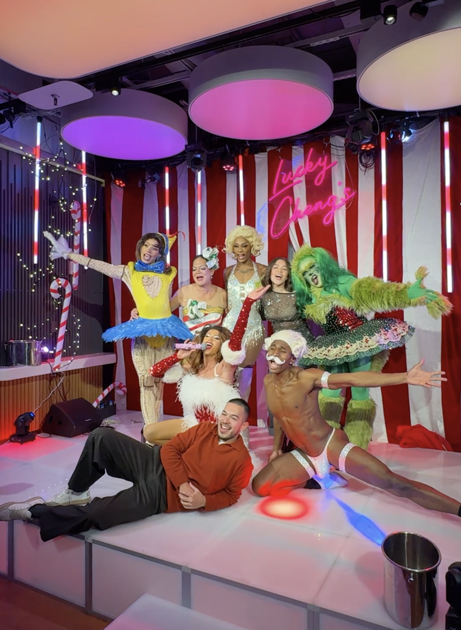 A group of drag performers and friends celebrating holiday attire and costumes on a decorated stage with a pink neon sign that reads 'Lucky Changs'.