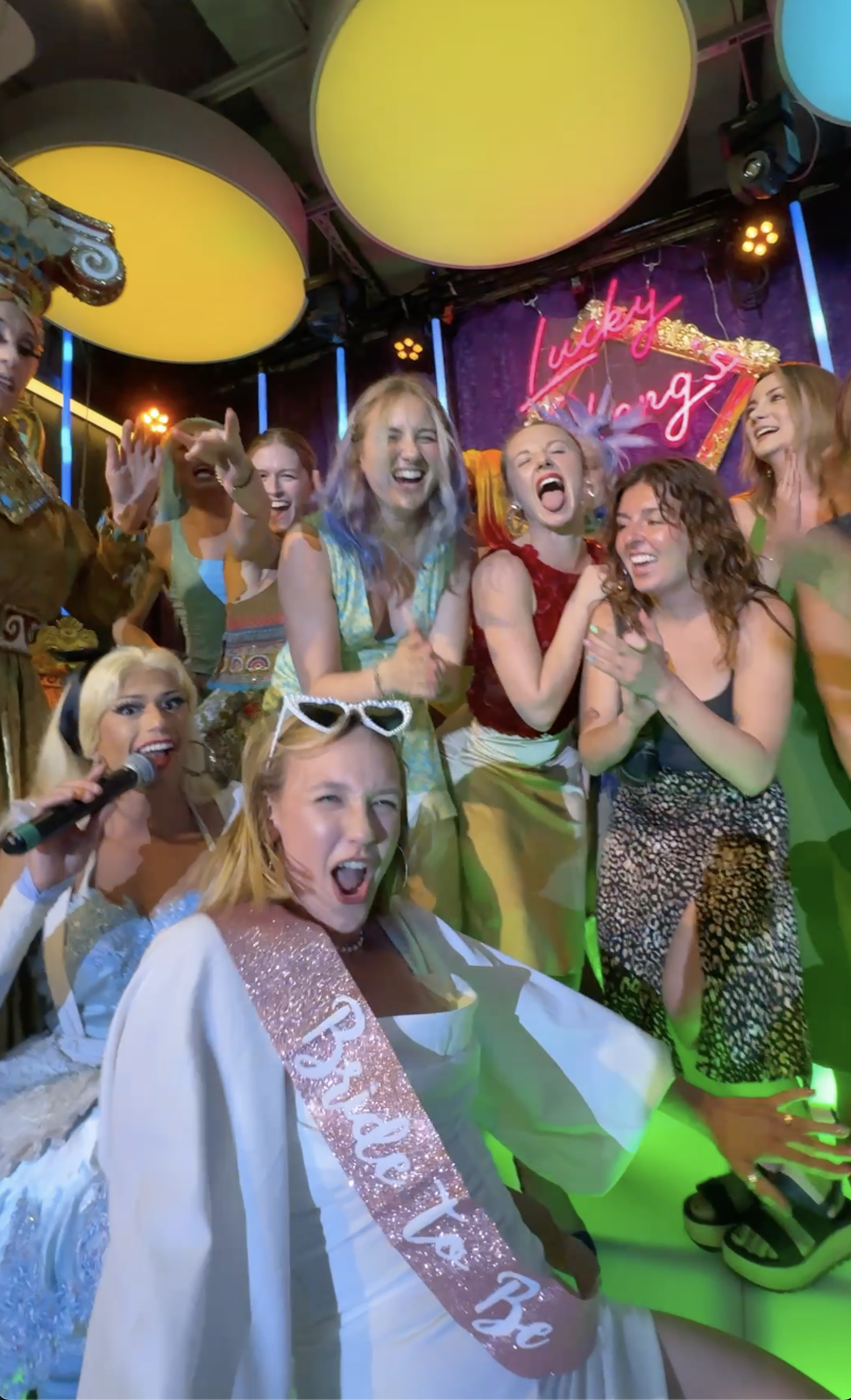 Group of women celebrating at a birthday party on a stage with colorful lighting and a neon sign that says 'Lucky Happy'. One woman is wearing a sash that says 'Birthday' and a tiara, and others are smiling and laughing.
