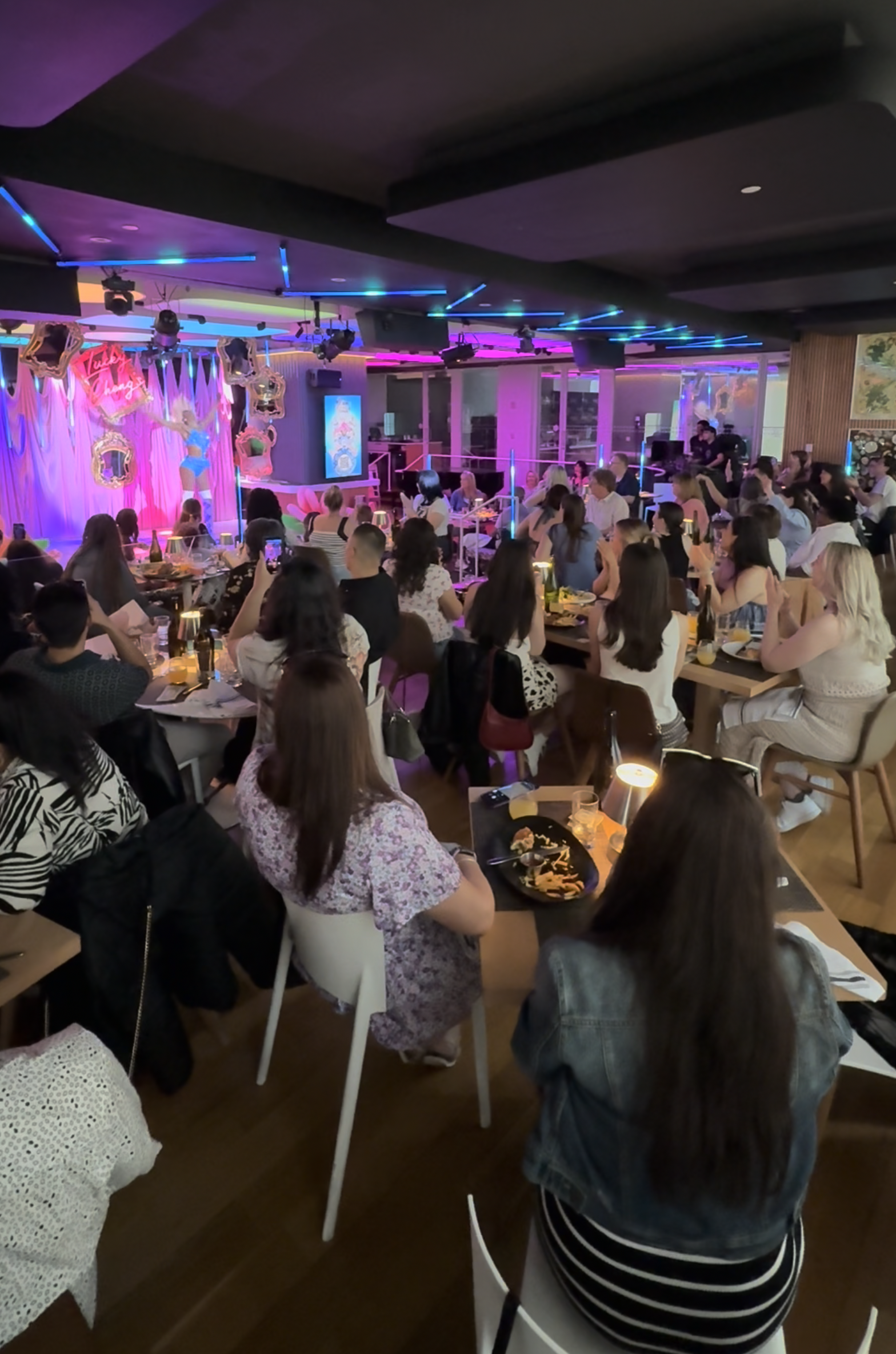 Crowd of people sitting at tables in a lively restaurant or bar with pink and blue neon lighting, decorated walls, and a stage area in the background.