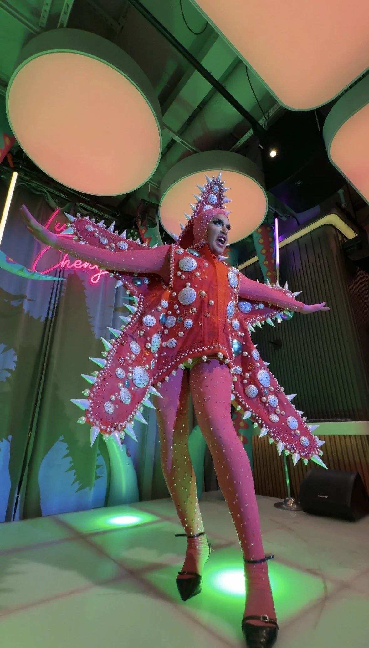 A performer on stage dressed in a bright pink, spiked, and decorated costume, with their arms spread wide, in front of green lighting and a pink neon sign. The costume is covered with large embellishments and spikes.