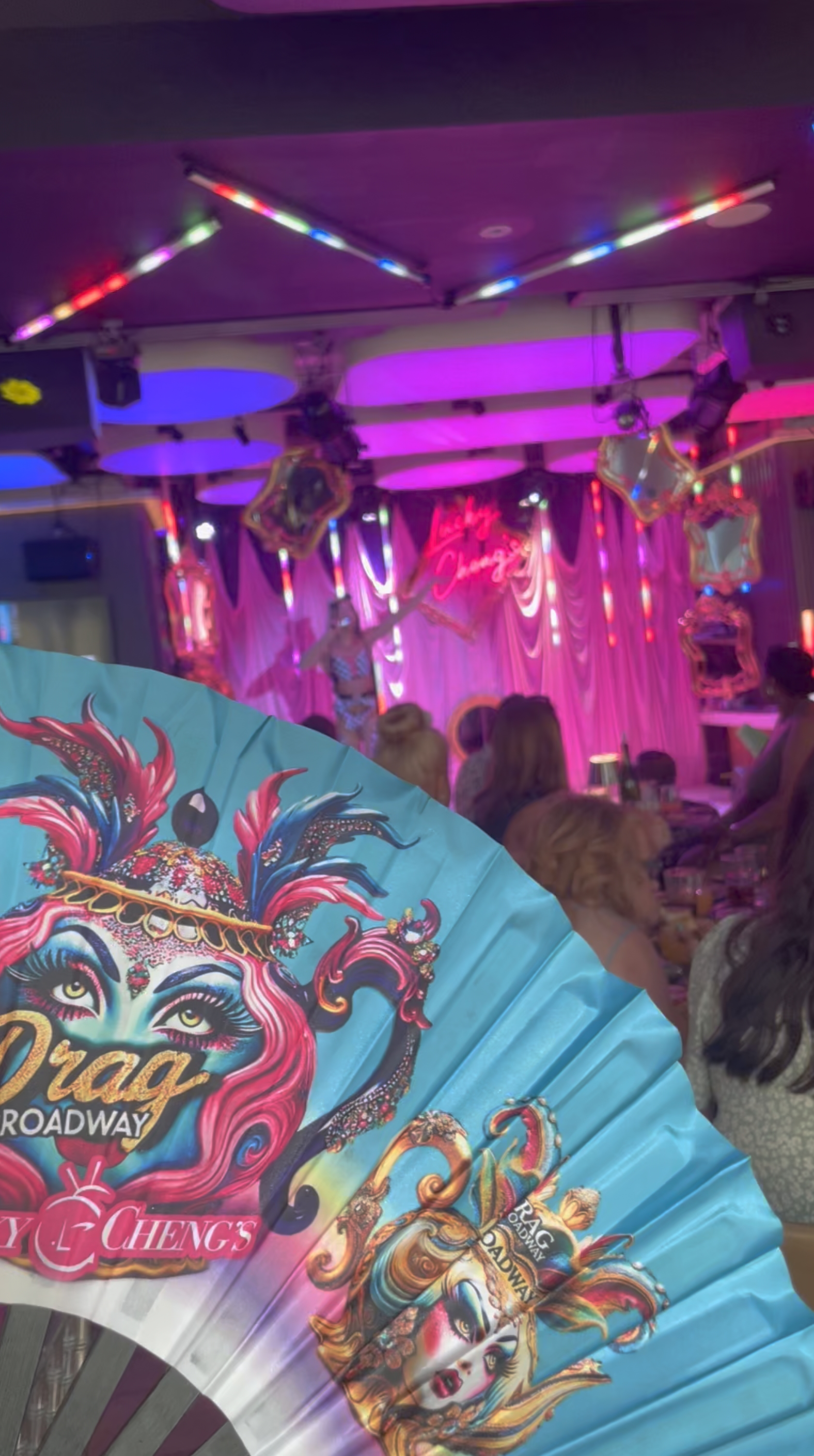Close-up of a colorful, illustrated promotional fan featuring a stylized woman with pink hair and theatrical makeup, advertising Drag Queen performances, with a stage and audience in the background decorated with pink and purple lighting.