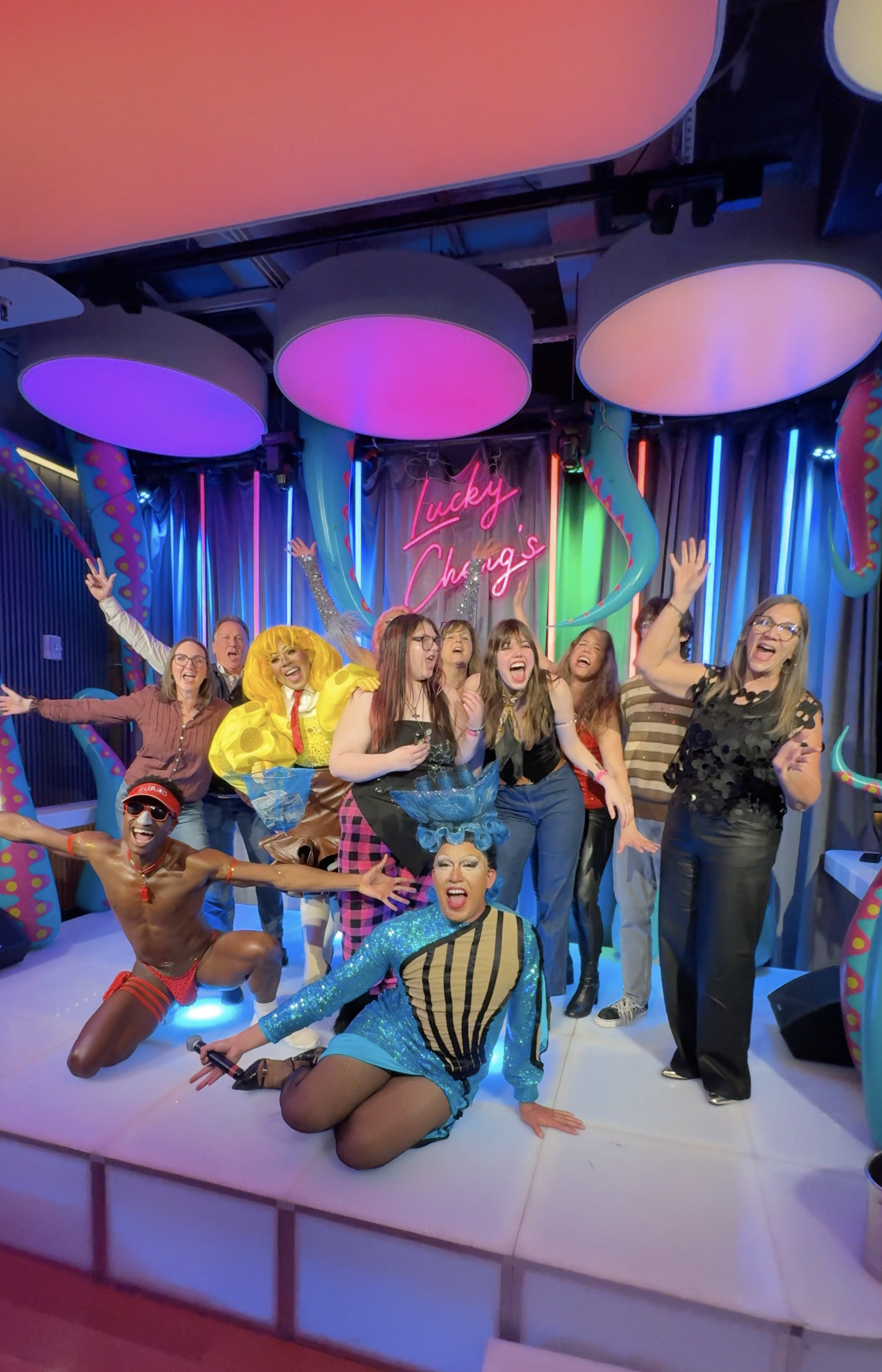 A group of people celebrating on stage with a colorful, neon-lit background and a sign that says 'lucky Charms'. The group includes individuals dressed in vibrant costumes, some dancing, laughing, and posing enthusiastically.