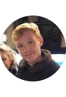 A young boy with reddish-blonde hair smiling at the camera, wearing a dark jacket, with part of a person visible next to him, inside a vehicle or similar setting.