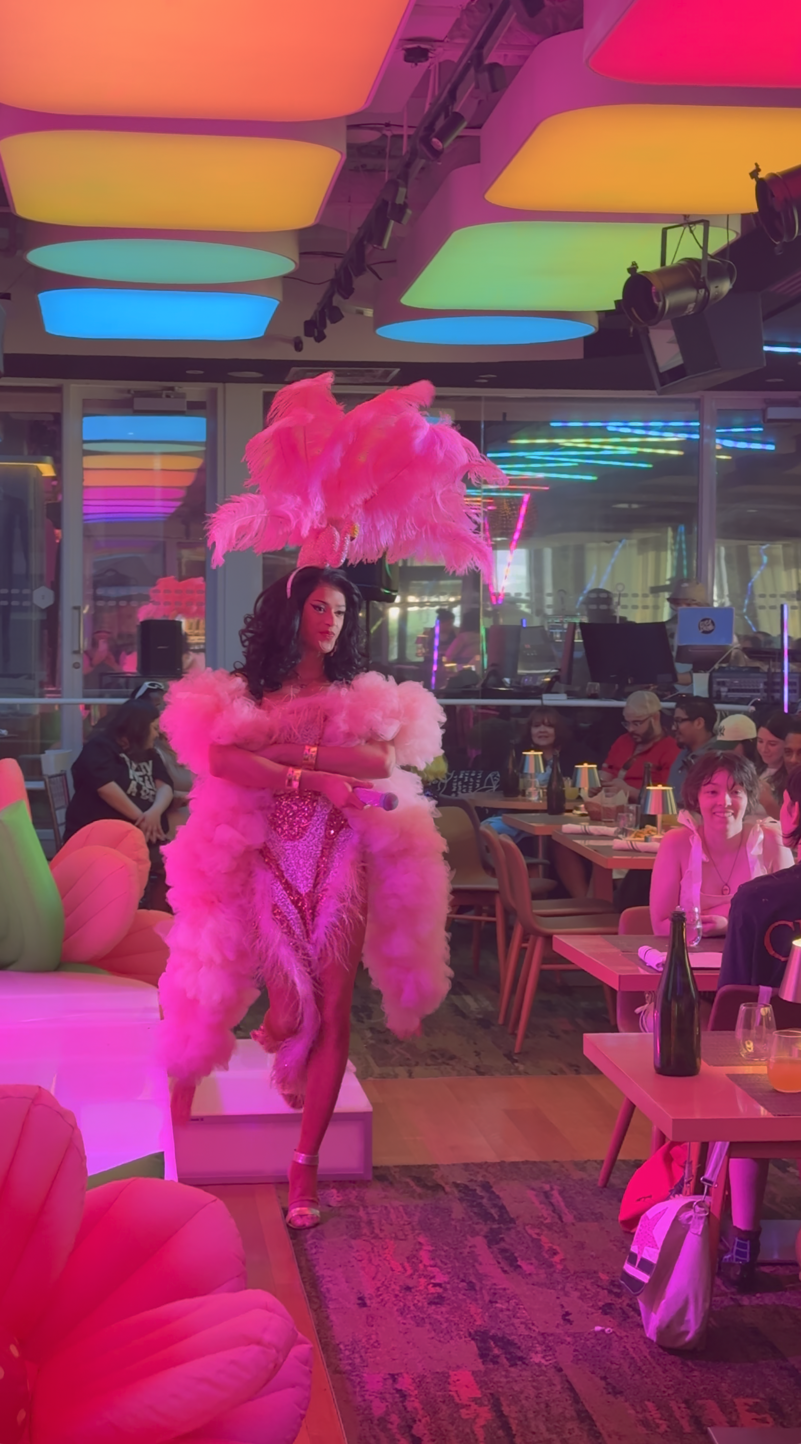 A performer wearing a vibrant pink feathered costume and high heels holding a microphone in a lively, colorful indoor venue with neon lights and an audience seated at tables.