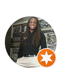 Smiling woman with long dreadlocks wearing a black top and beige pants, standing in front of comic book posters.