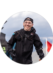 Smiling man in black outdoor gear, wearing a cap backward, standing outdoors with cloudy sky background.