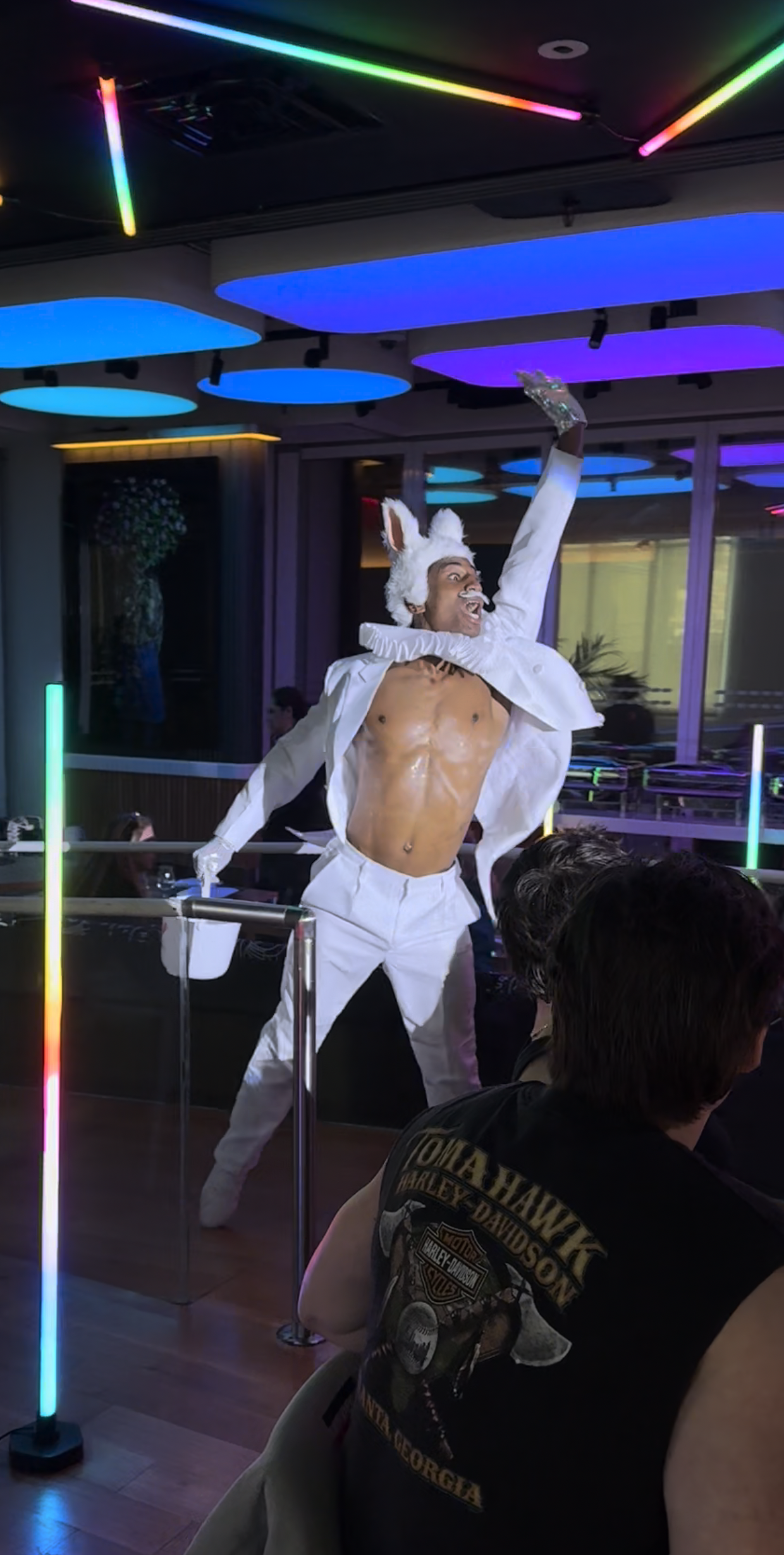 Performer dressed as a rabbit with ears, white gloves, and a ruffled collar, dancing on stage in a nightclub with colorful neon lighting.
