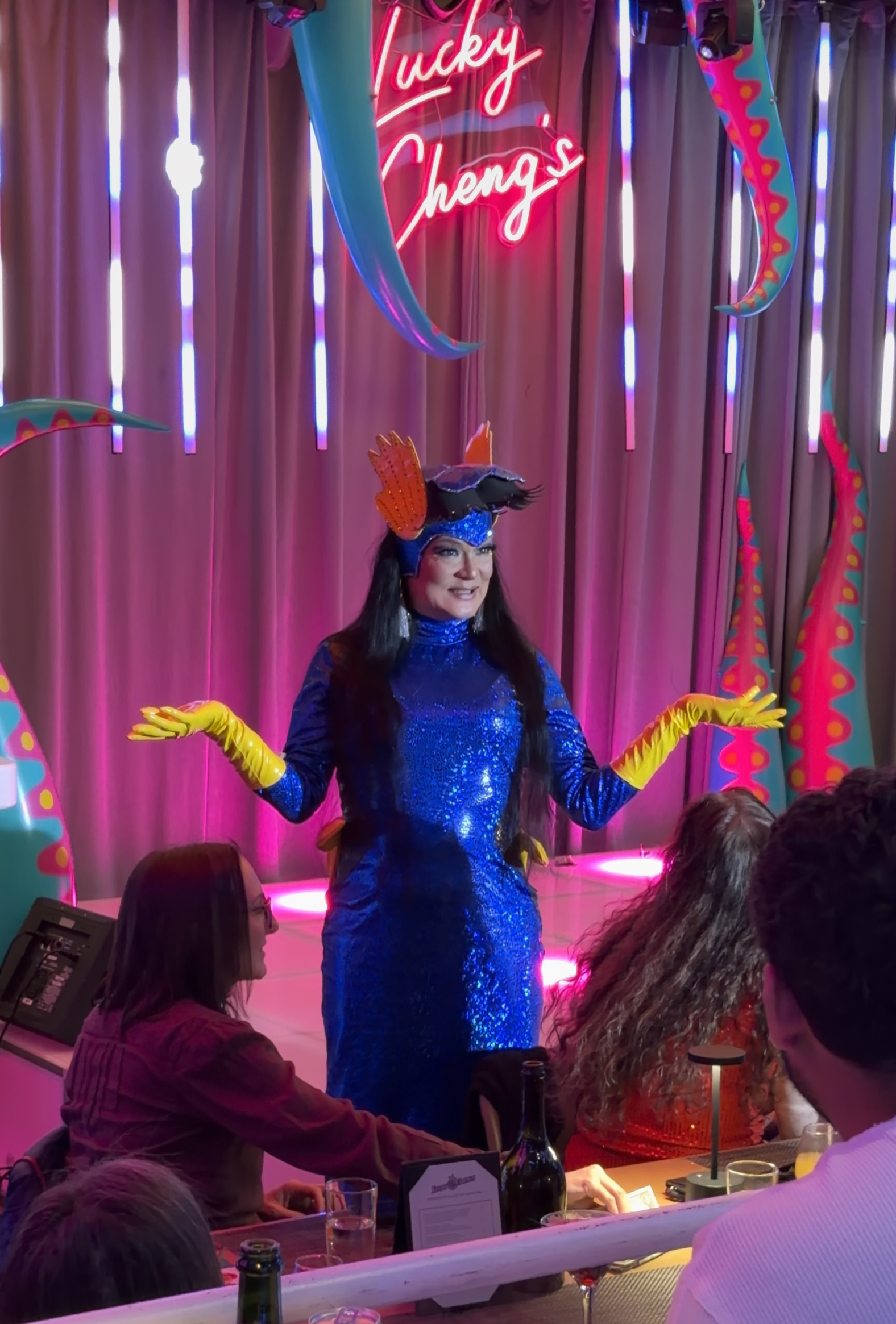 A woman dressed in a blue sequined gown and yellow gloves is performing on a stage decorated with colorful horns and a pink curtain. She is wearing a fish-shaped headpiece and smiling. A pink neon sign above her reads 'Lucky Cheng's'.
