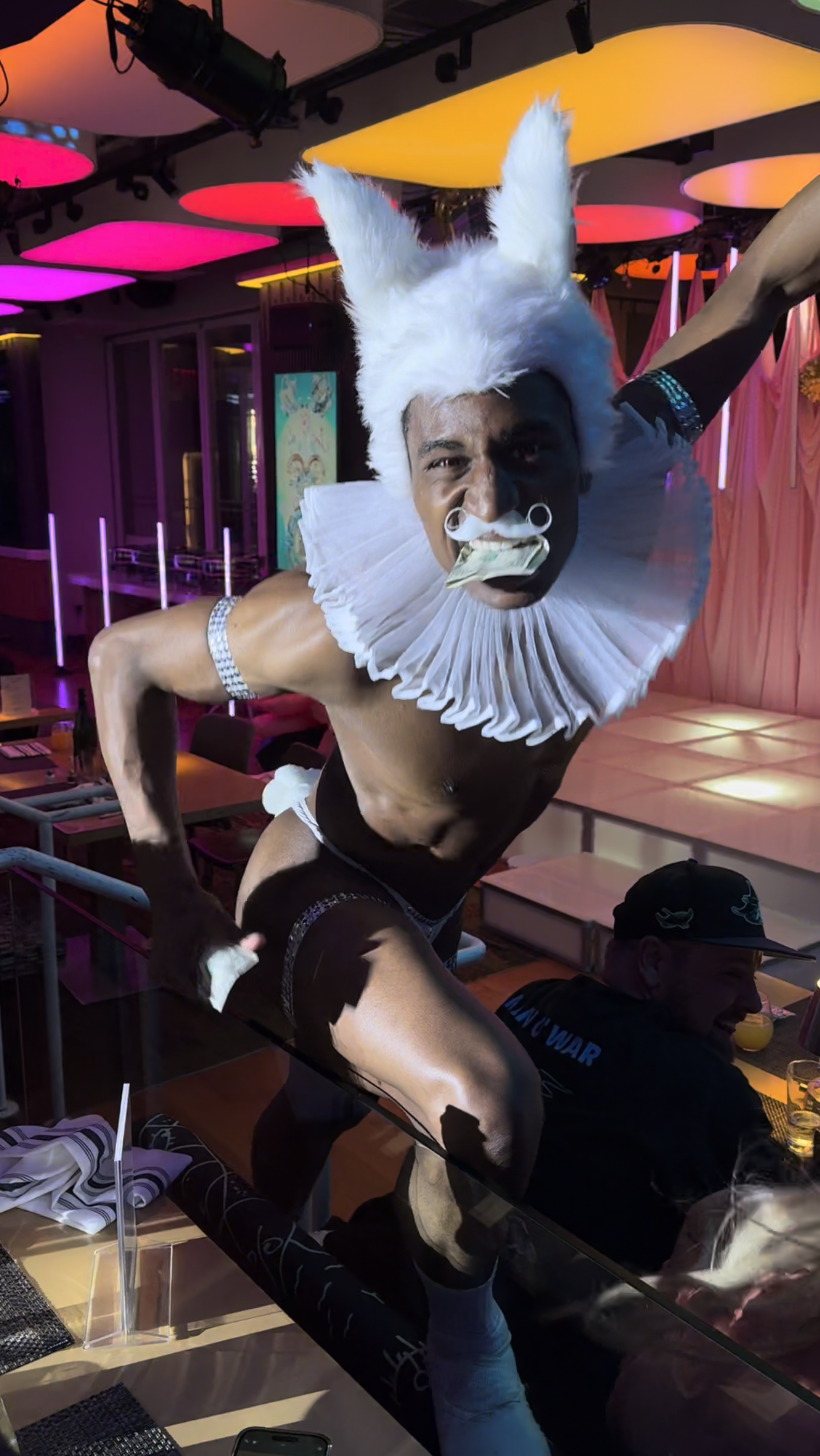 A person dressed as a clown with a white wig, mustache, ruffled collar, and minimal clothing is performing a daring pose on a glass table in a colorful venue with pink, purple, and yellow lighting.