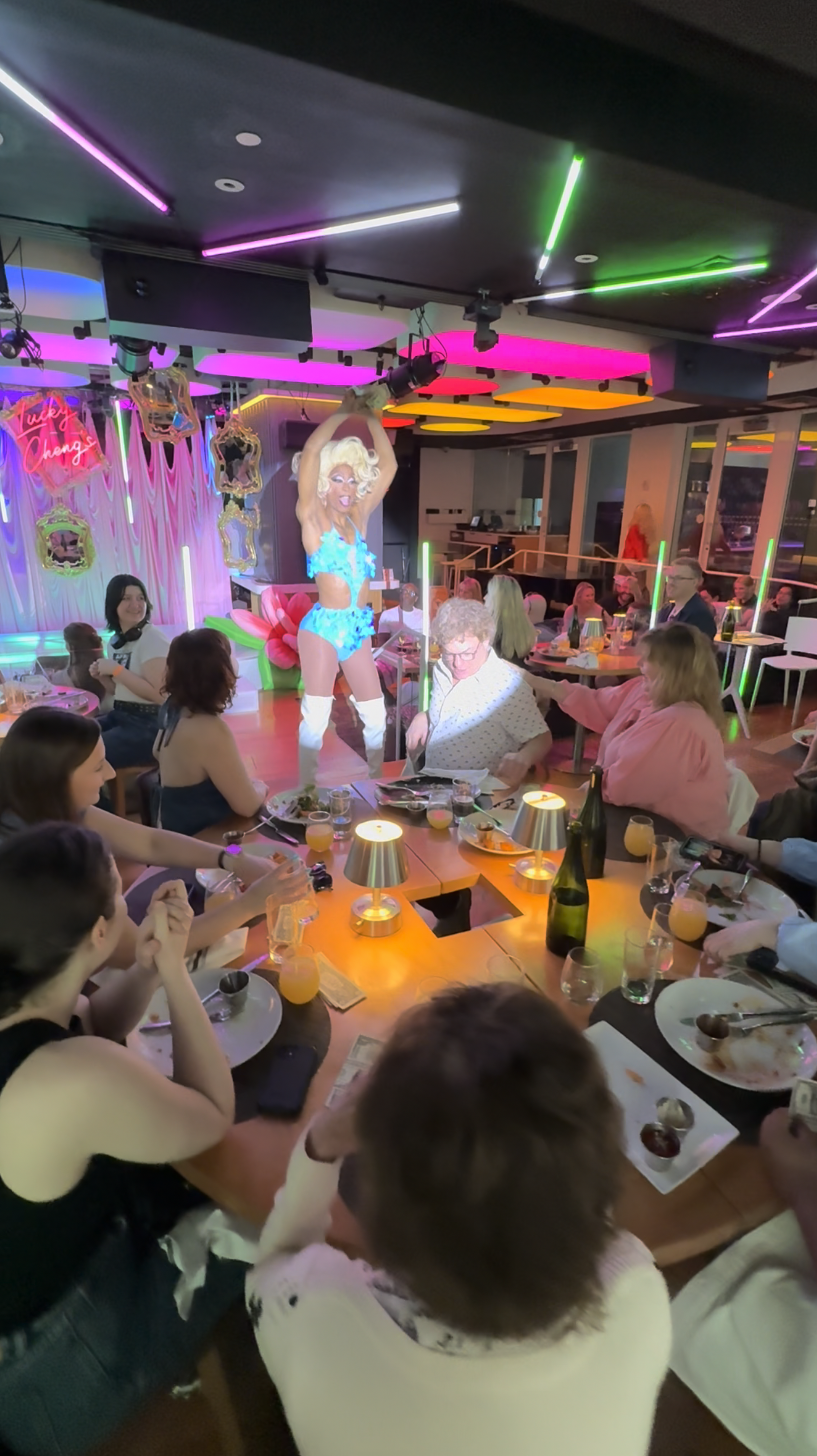 A lively restaurant or club scene with a performer on stage dressed in a bright blue costume, entertaining seated guests. The room is decorated with colorful neon lights and ornaments, creating a vibrant atmosphere.