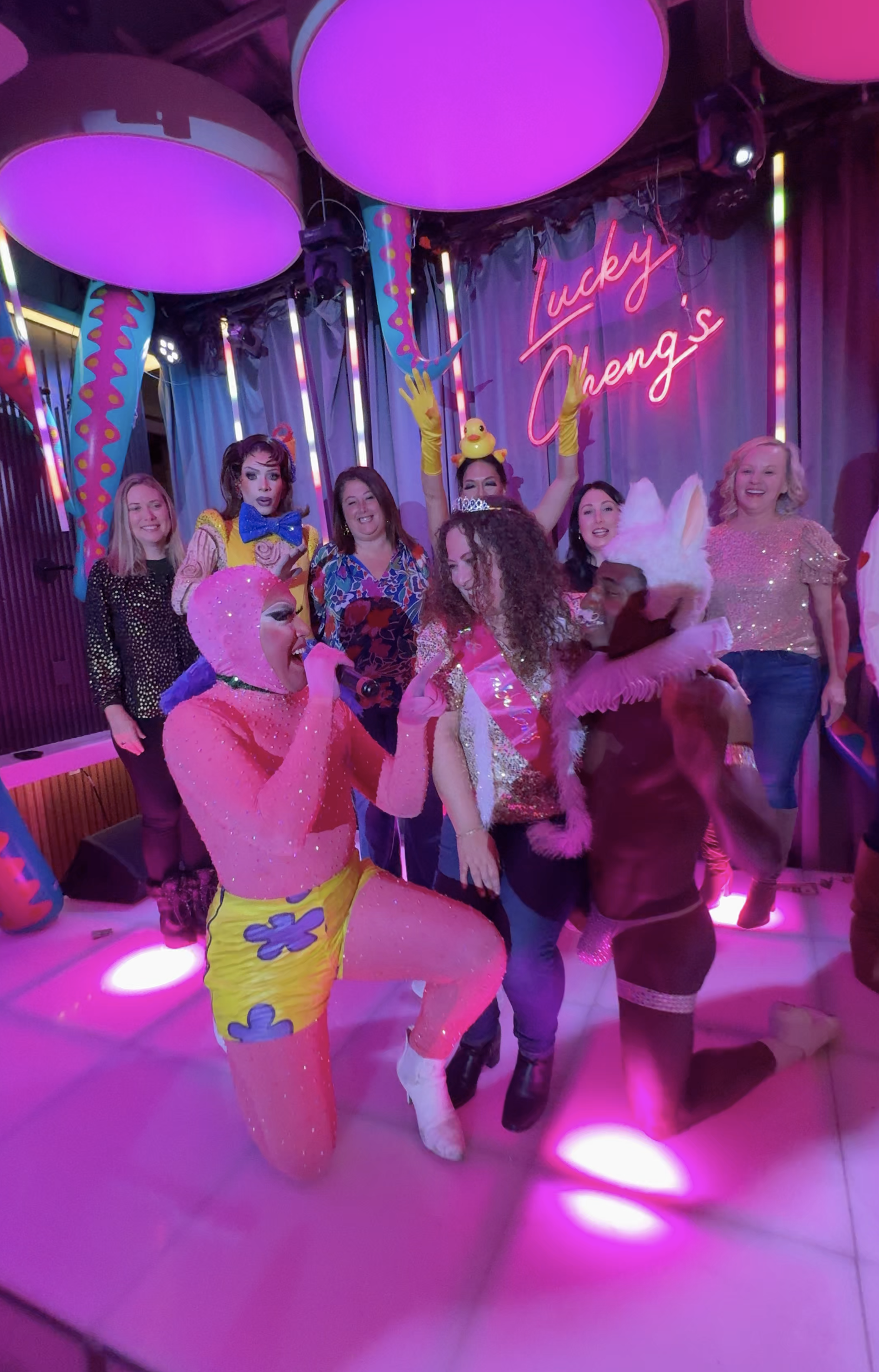 A group of people in colorful costumes celebrating at a party with neon lights, including a person dressed as a clown and another in bunny ears, in front of a sign that reads 'Lucky Cheng's'.