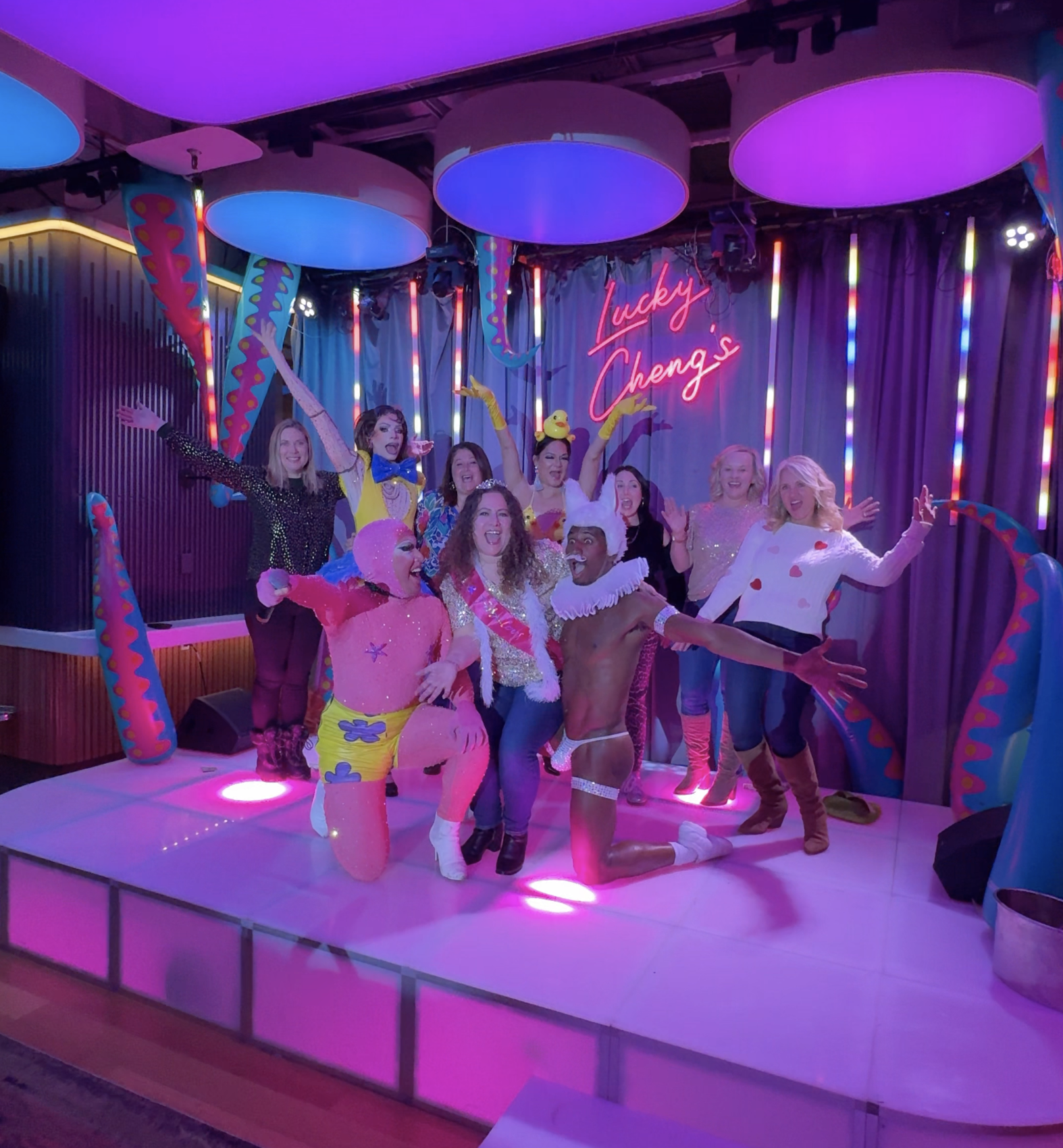 Group of performers dressed in colorful costumes and makeup posing on stage with a pink neon sign that reads 'Lucky Cheng's', decorated with bright neon lights and large hanging soft circle decor.