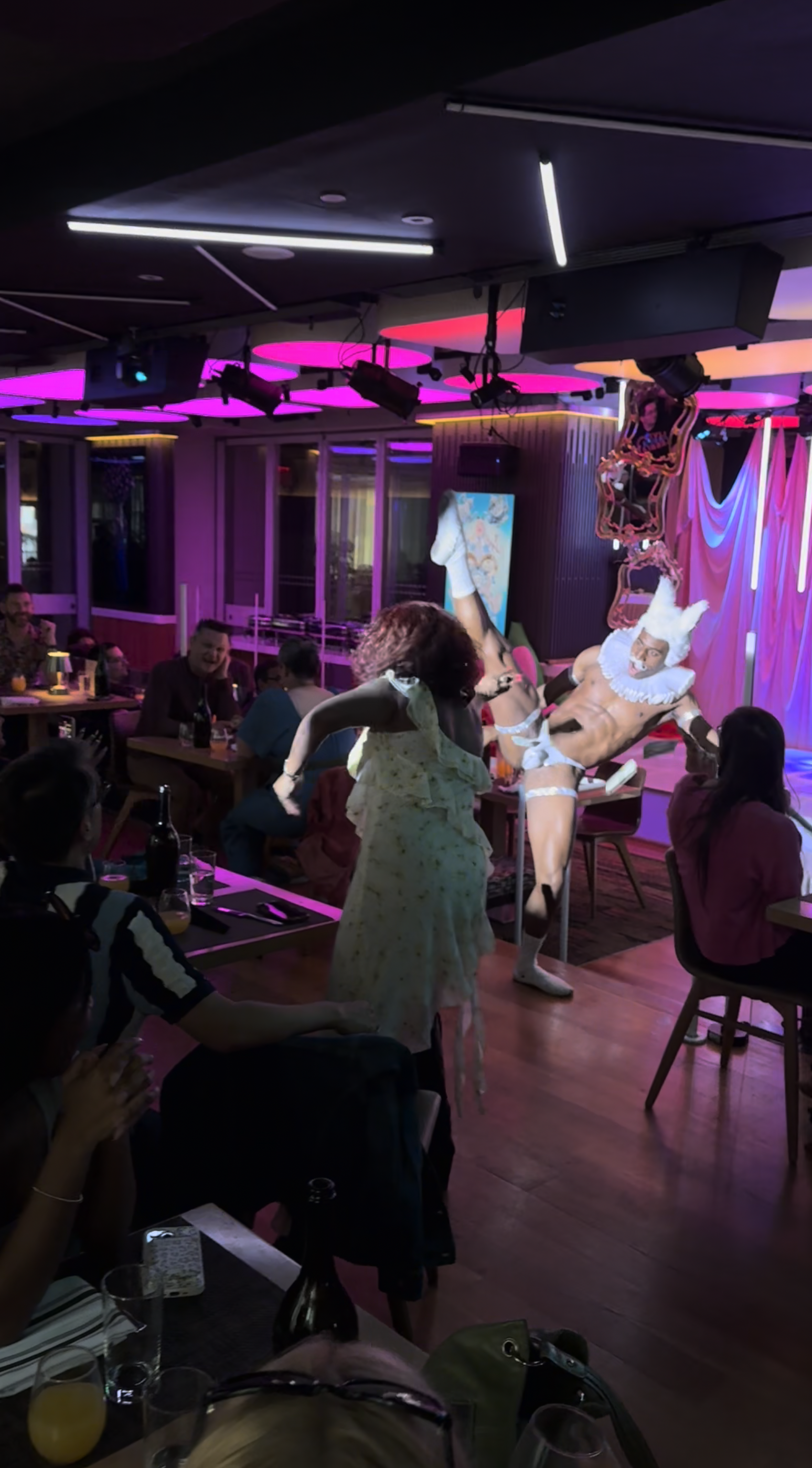 A lively indoor scene at a nightclub or bar where a performer dressed as a clown is entertaining a seated audience. The performer is wearing a white curly wig and makeup, and is in a dynamic pose. The setting has colorful lighting with pink and purpl