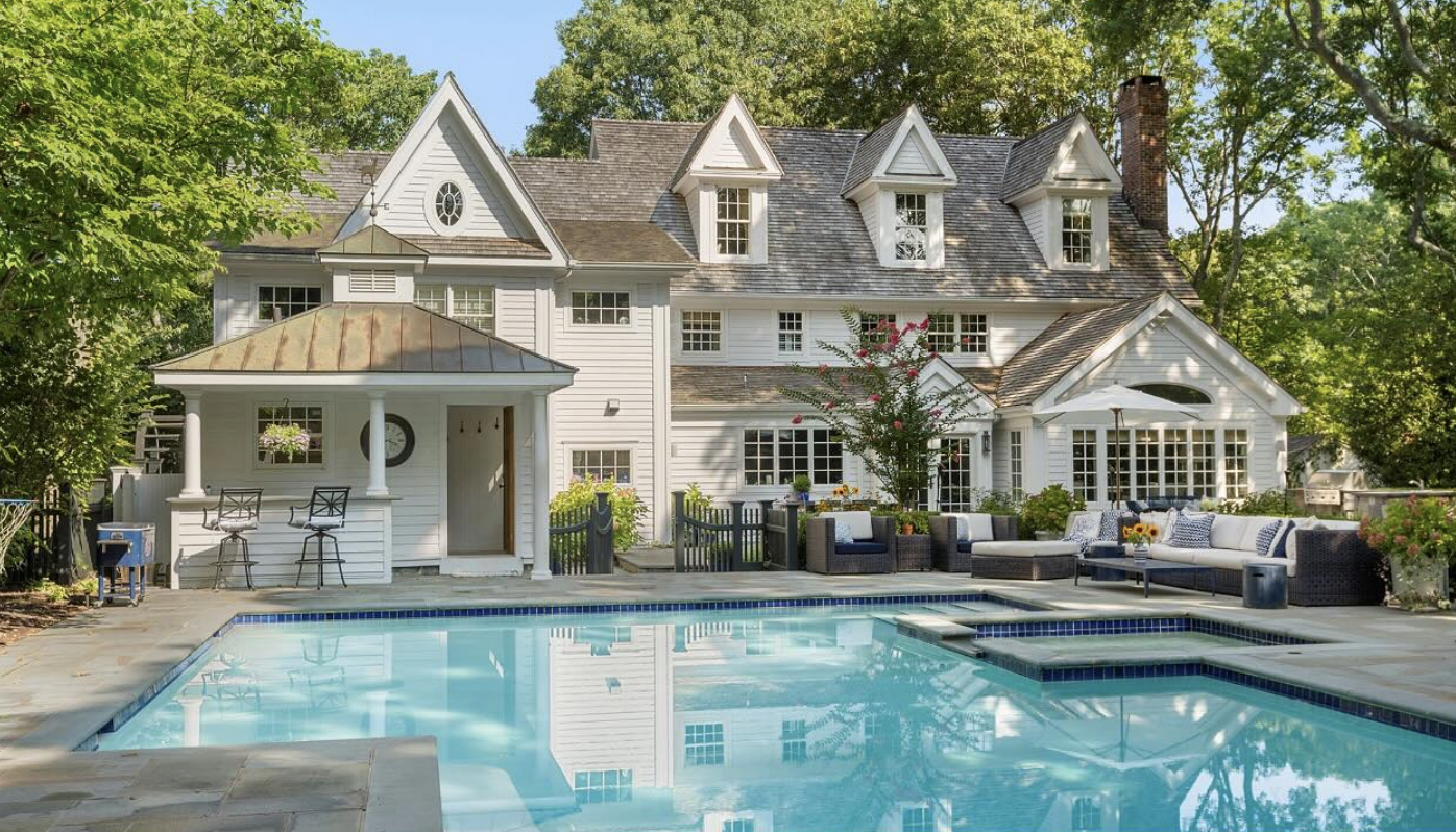 White multi-story house with a swimming pool in the backyard, surrounded by greenery and outdoor furniture, including cushioned sofas and a small pergola.