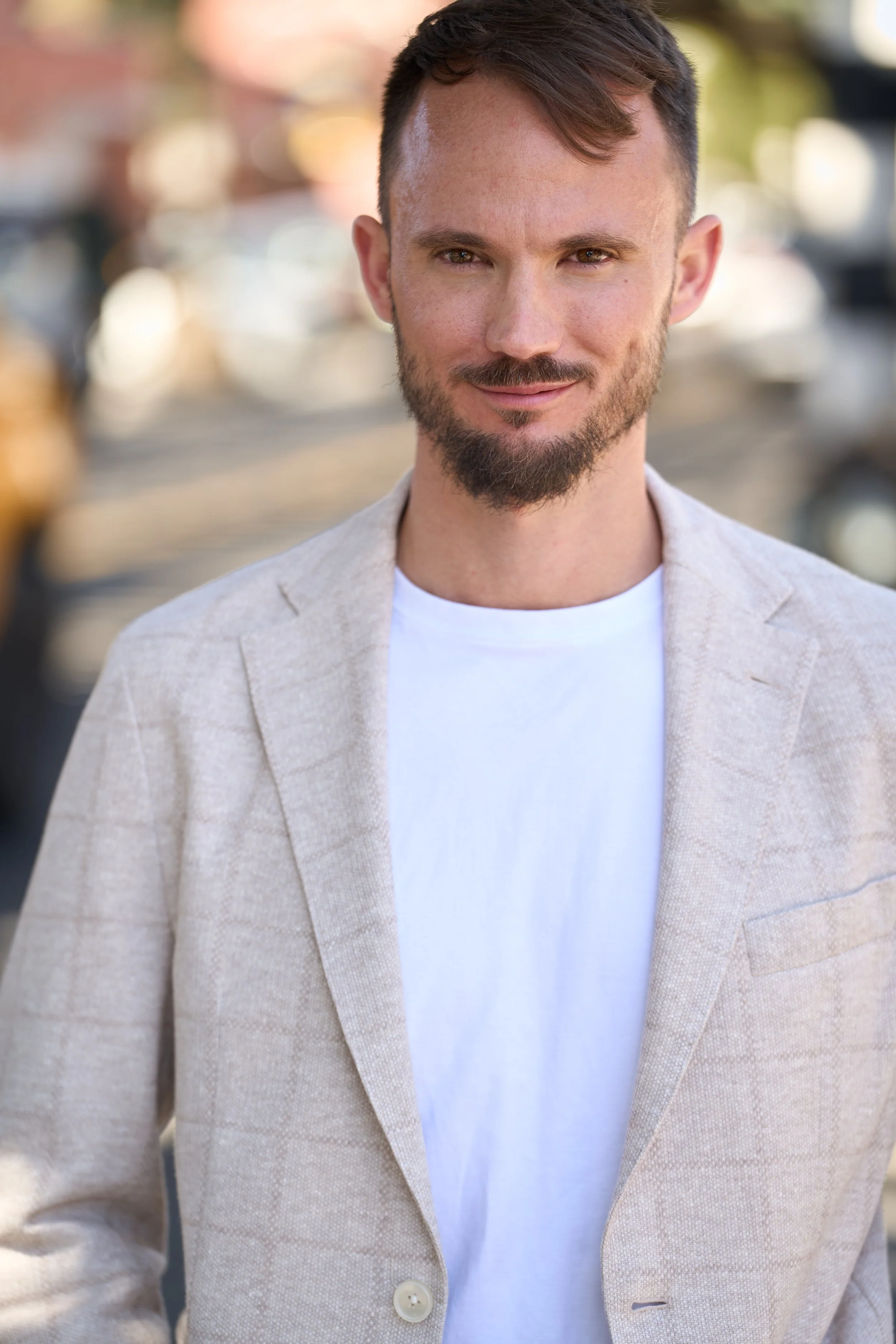 Close-up portrait of a young man with short brown hair and a beard, wearing a light beige blazer and a white T-shirt, standing outdoors with a blurred background of trees and buildings.