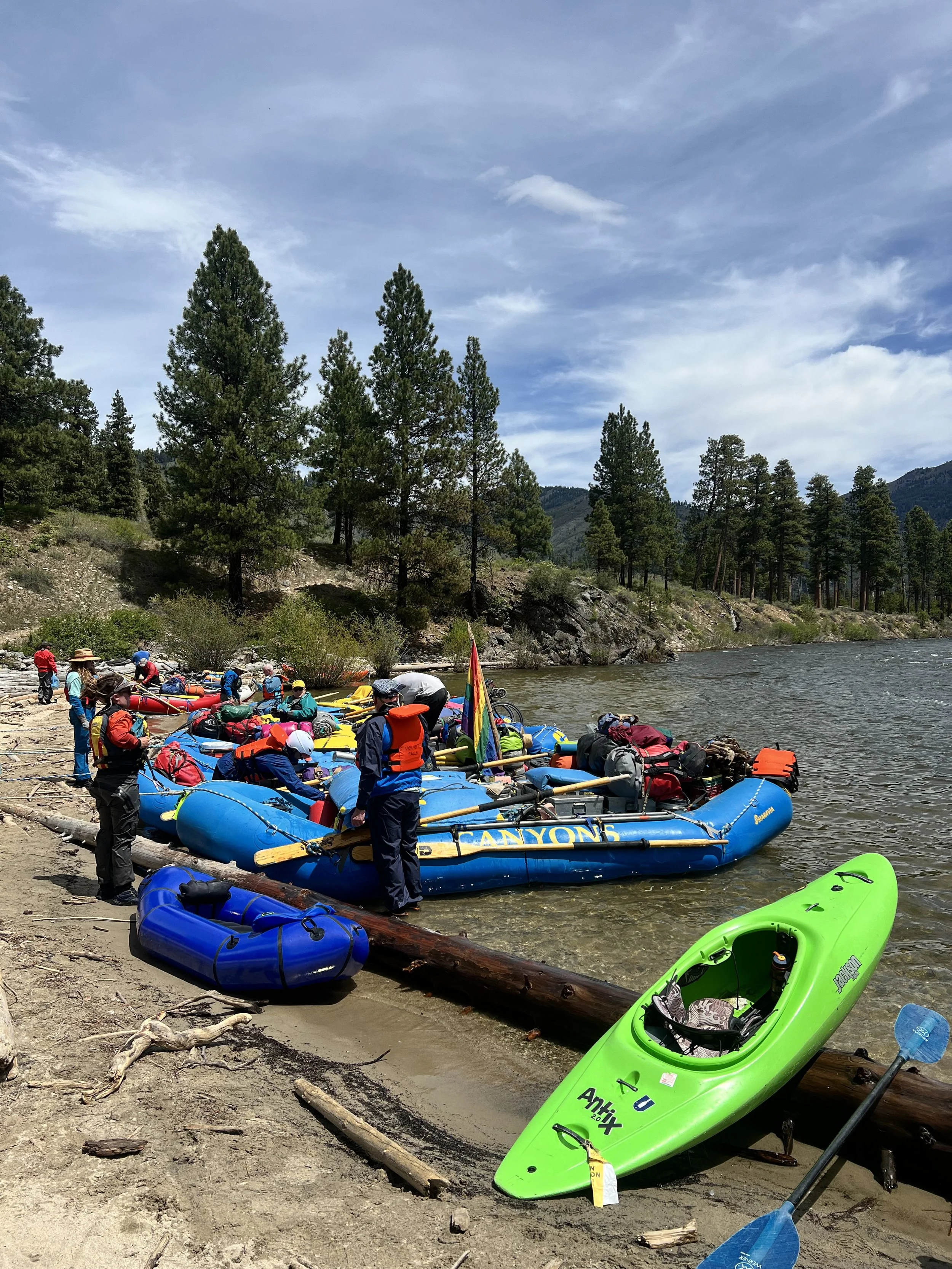 People preparing for river rafting with inflatable boats and paddles on a sandy riverbank surrounded by trees and mountains under a partly cloudy sky.