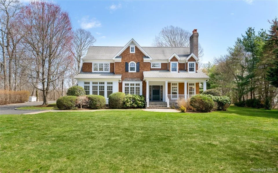 Large two-story house with brick and white siding, front porch, surrounded by a well-manicured lawn, bushes, and trees.