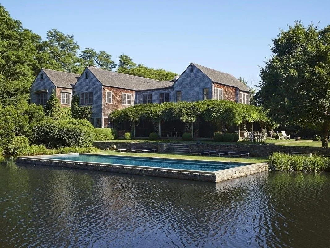 A large house with gray and brick exterior, large windows, and multiple gabled roofs, surrounded by lush green trees, with a swimming pool in the foreground, and a waterfront view.