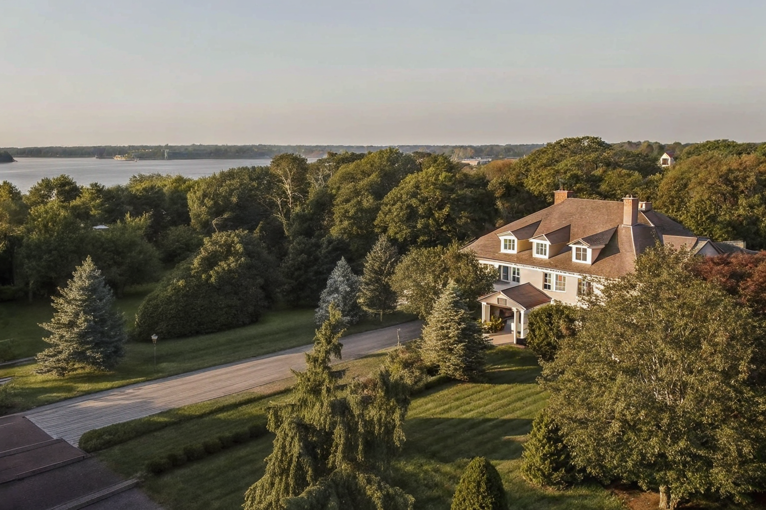 A large house surrounded by trees and greenery, with a view of a river or lake in the background during sunset.