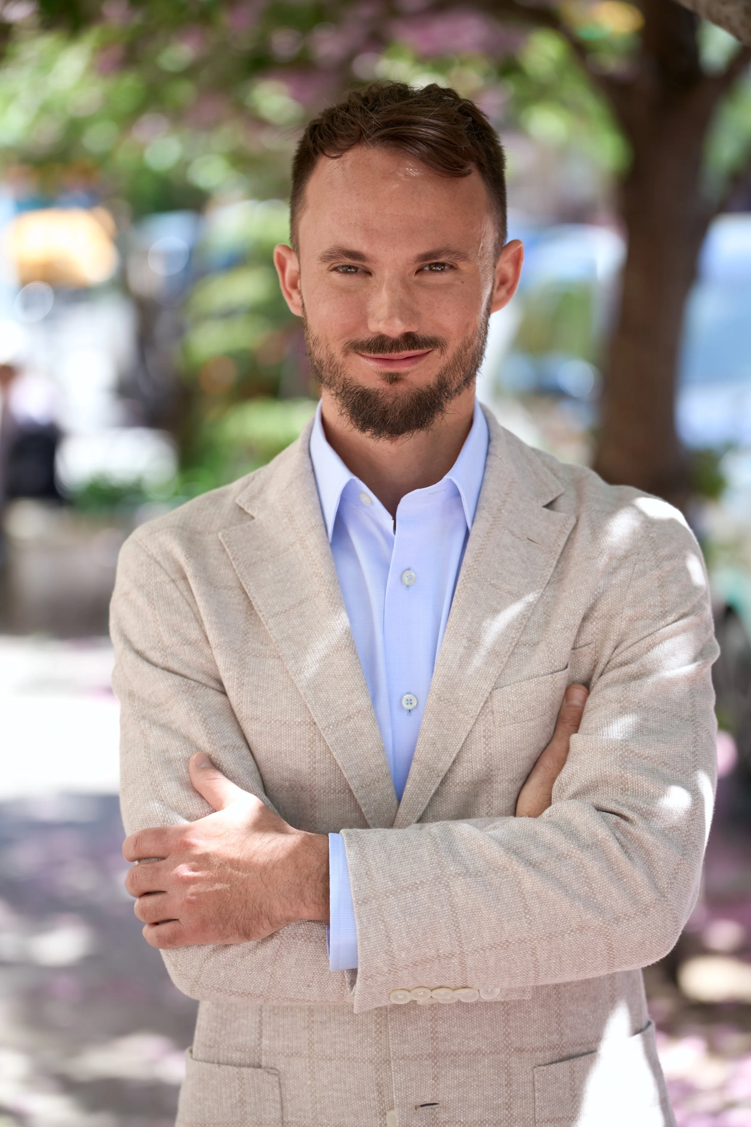 A man wearing a light-colored suit with a white shirt, standing outdoors with trees and blurred background, smiling with arms crossed.