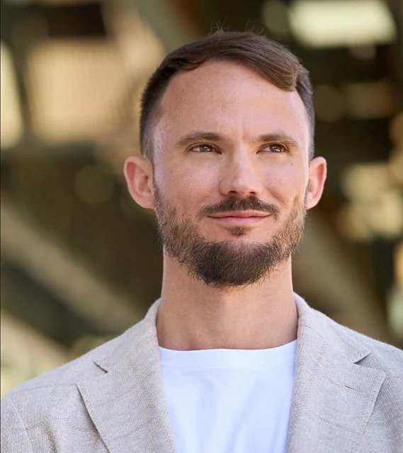 A man with short brown hair and a beard, wearing a light gray blazer and a white t-shirt, looking thoughtfully to the side outdoors.
