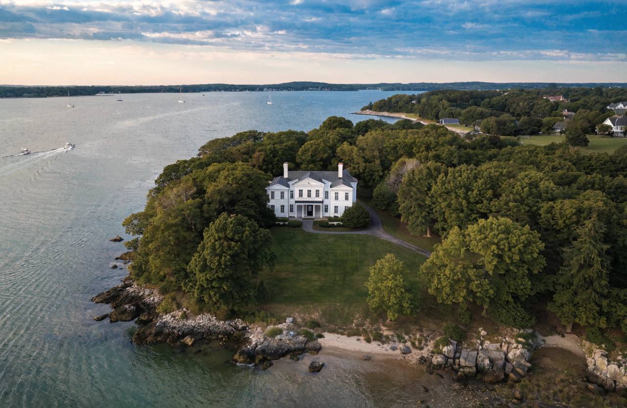 A large white mansion on a rocky shoreline surrounded by green trees, facing a body of water with sailboats, under a partly cloudy sky.