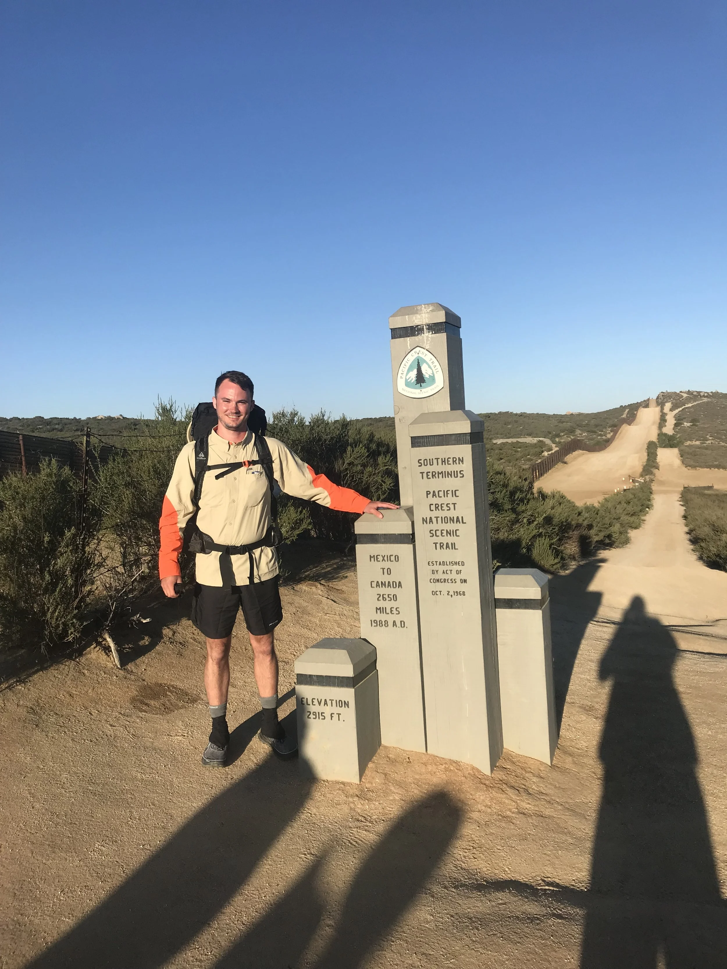 A man standing next to a stone marker at the Southern Terminus of the Pacific Crest National Scenic Trail, with a dirt trail extending into the distance and a clear blue sky above.