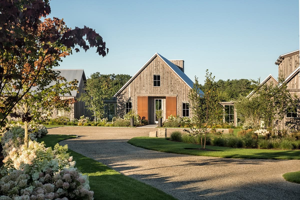 A peaceful rural scene featuring a modern farmhouse with wood siding, surrounded by manicured lawns, trees, and flowering plants, with a gravel driveway leading to the house.
