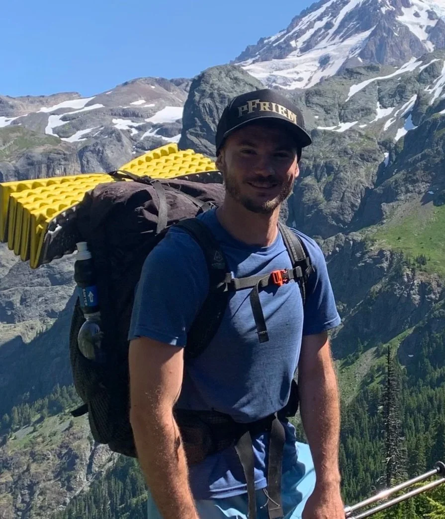 Man hiking in the mountains with snow-capped peaks, green forests, and rocky terrain in the background, wearing a cap, backpack, and outdoor clothing.