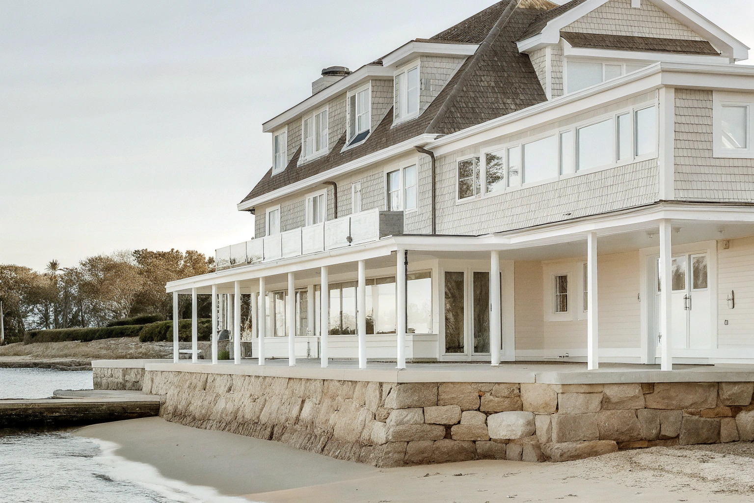 Beachfront house with a stone foundation, white wooden porch, and multiple windows overlooking the water, with trees in the background.