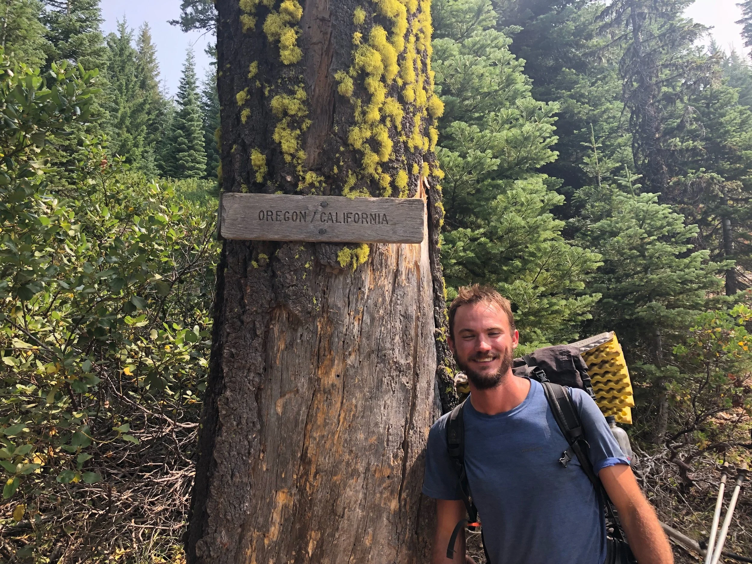 A man with a backpack smiling in front of a tree with a wooden sign that reads "Oregon / California" in a forested area with green trees and foliage.