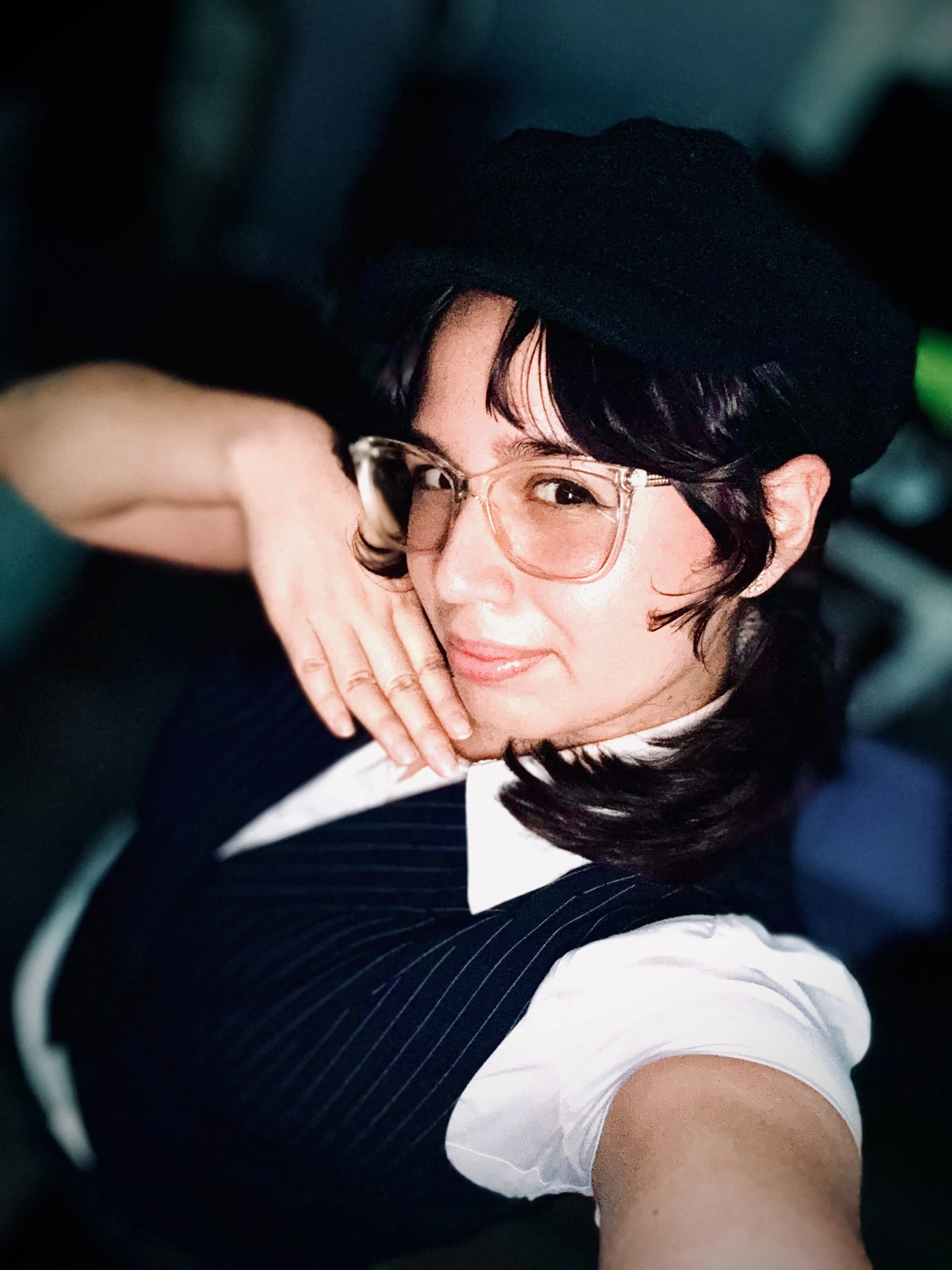 A person with glasses, black hair, wearing a black beret and a striped suit, smiling and resting their head on their hand.