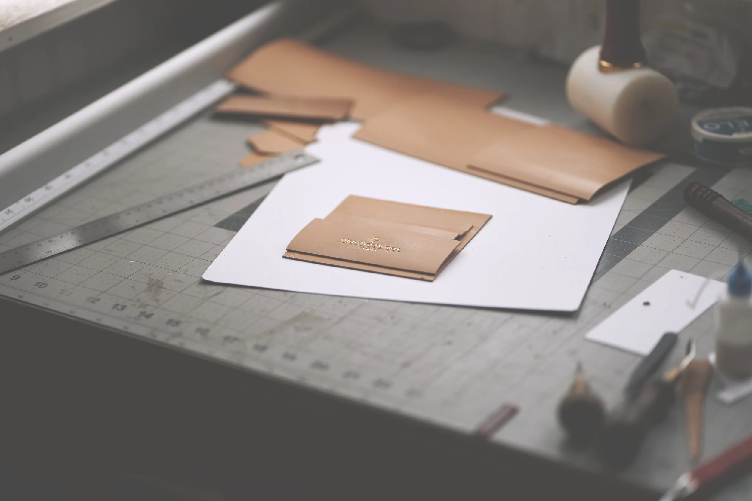 A workspace with cut pieces of tan leather, a metal ruler, a white sheet of paper, and crafting tools including a roller and a marker, on a cutting mat.