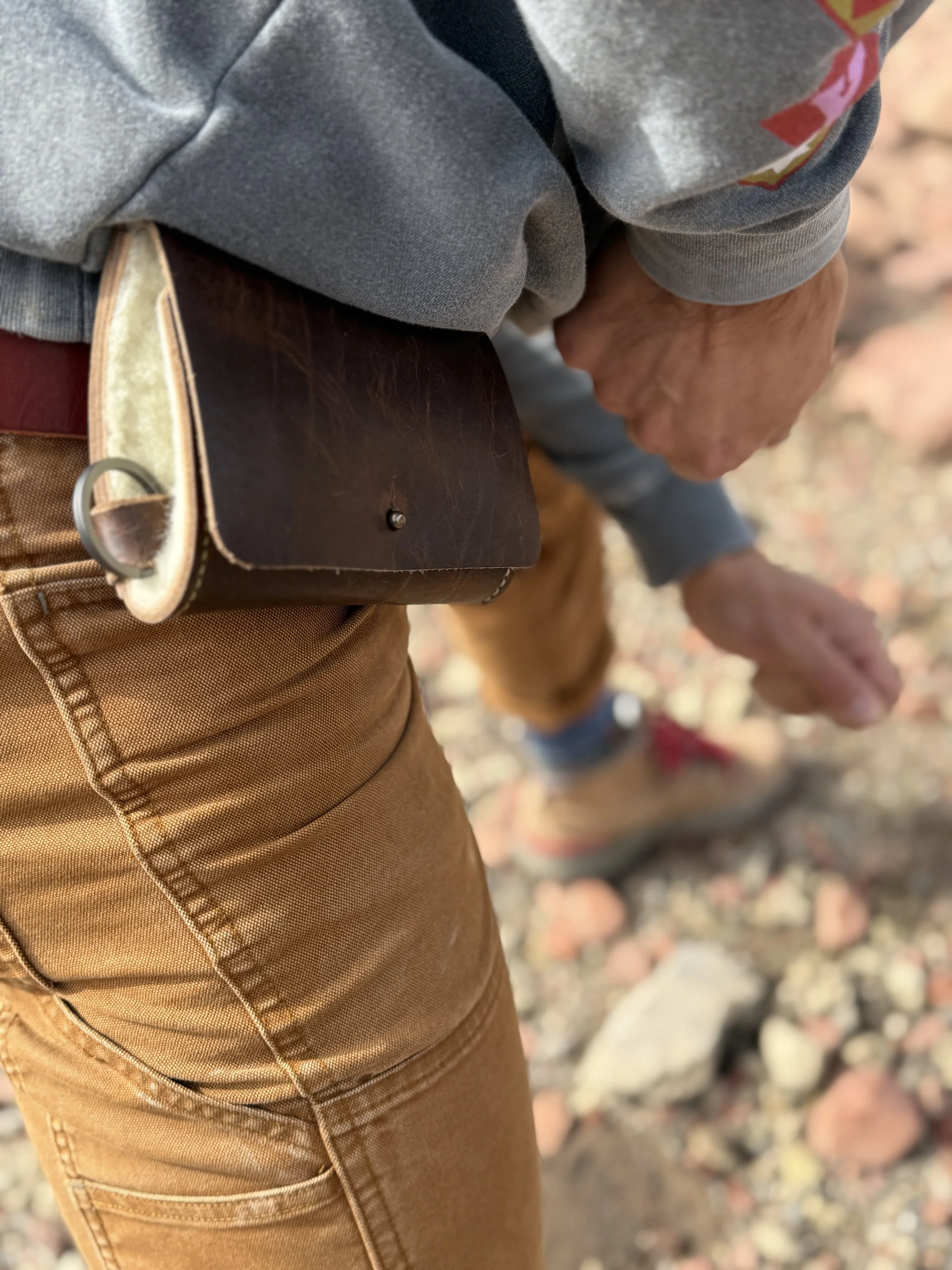 Close-up image showing a person wearing a gray sweatshirt and tan pants with a brown leather belt. The person's hand is resting on their thigh, and the background appears to be rocky or gravelly terrain.