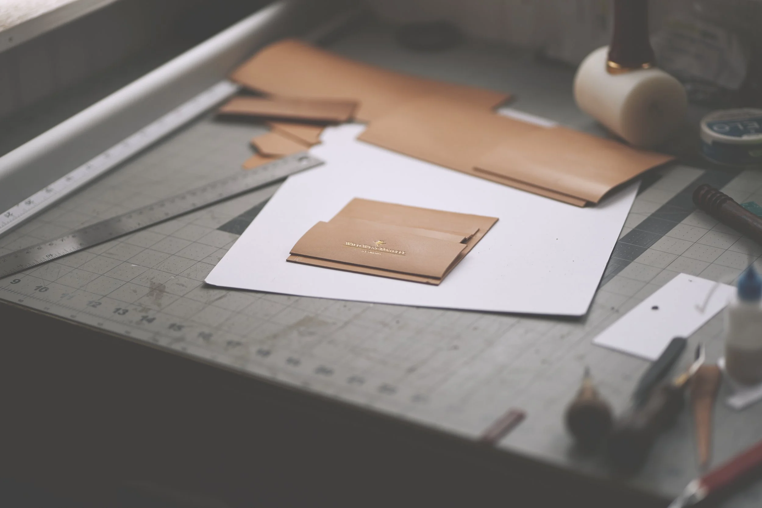 Close-up of a worktable with leather pieces, a ruler, a metal tool, a paper, a roller, and other tools used for leatherworking.