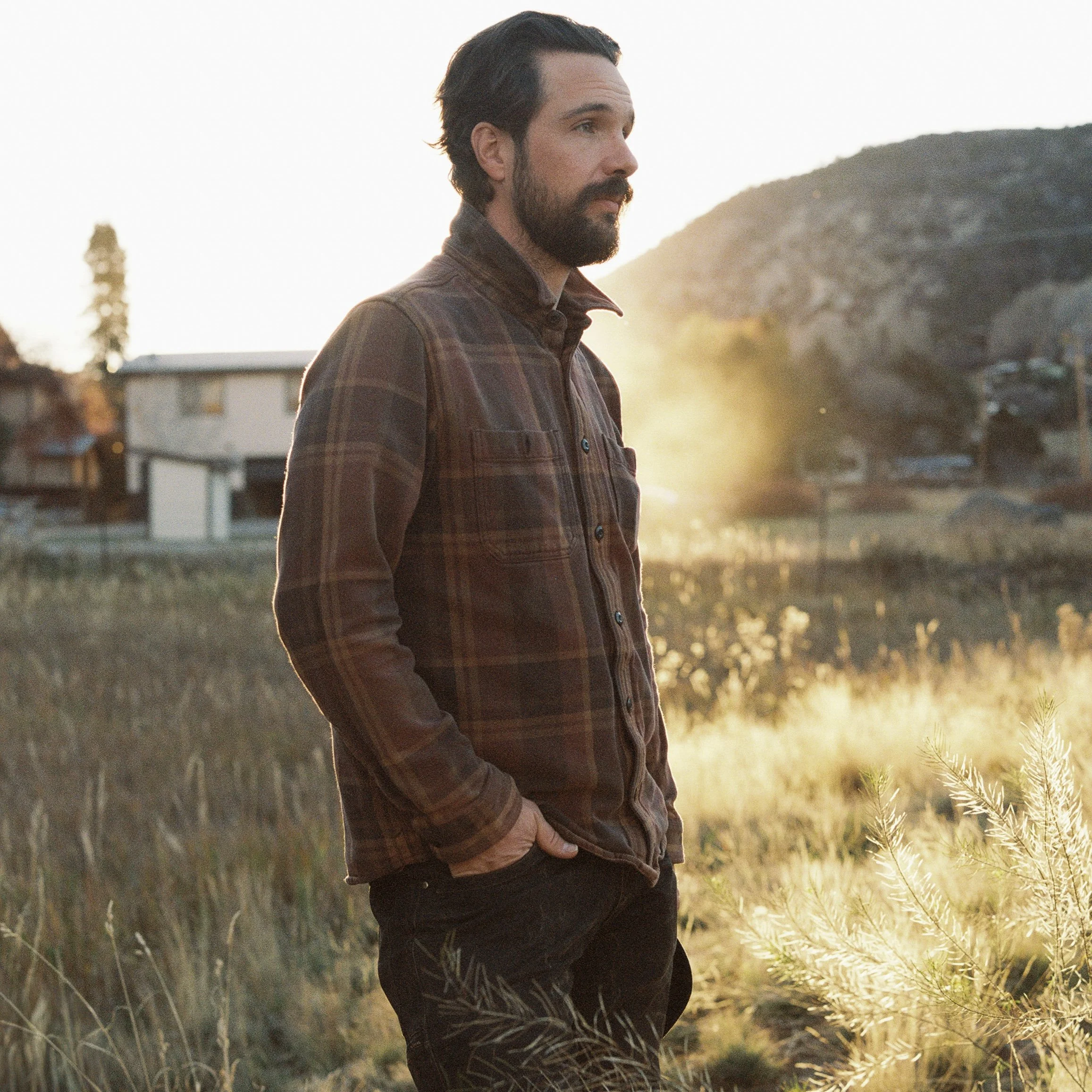 A man with a beard and dark hair standing outdoors in a field at sunset, wearing a plaid jacket and looking to the side.
