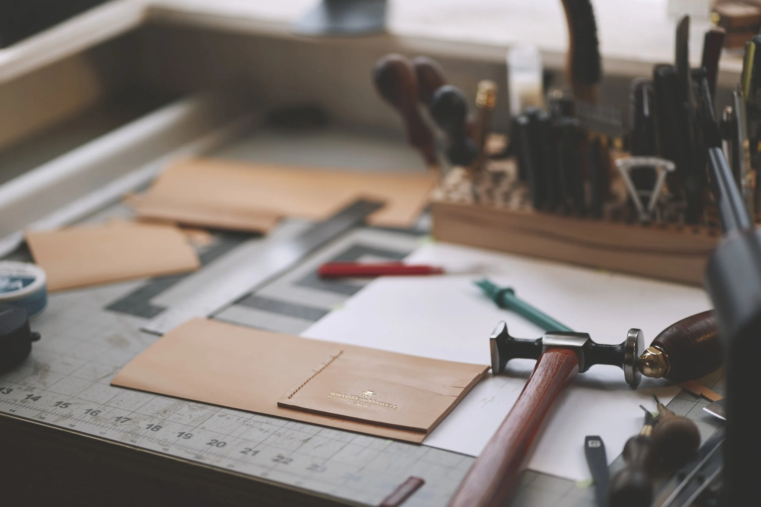 Workstation with leather samples, tools, and drafting supplies, including featuring a leather swatch with the printed logo 'Vera City Leather' and a ruler on the table.