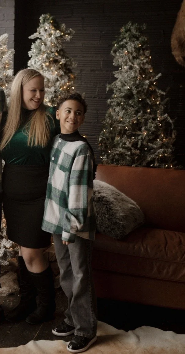 Two people standing next to Christmas trees decorated with lights, in front of a dark stone wall, in a cozy indoor setting.
