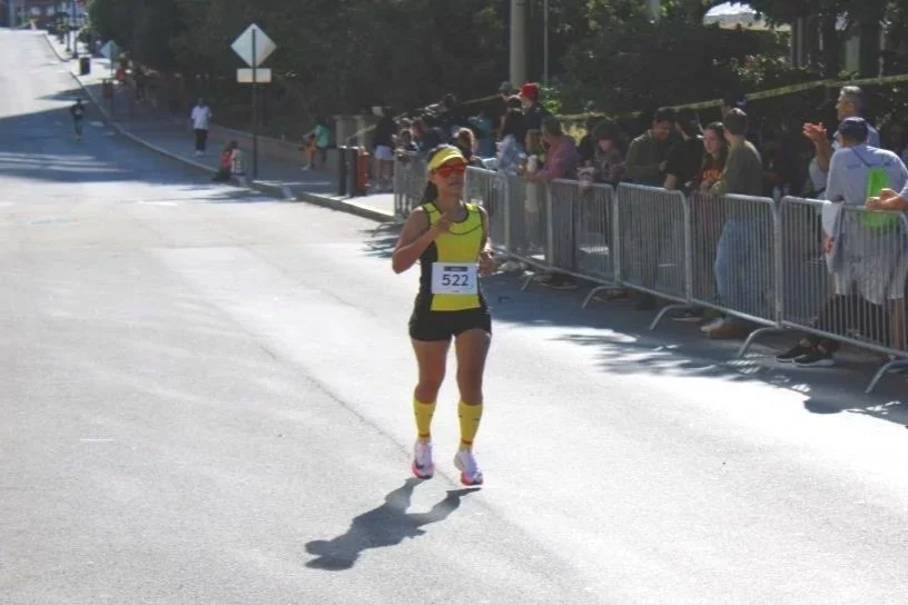 Female runner wearing a yellow athletic outfit participating in a race on a sunny day, with spectators behind a metal barricade on the side of the street.