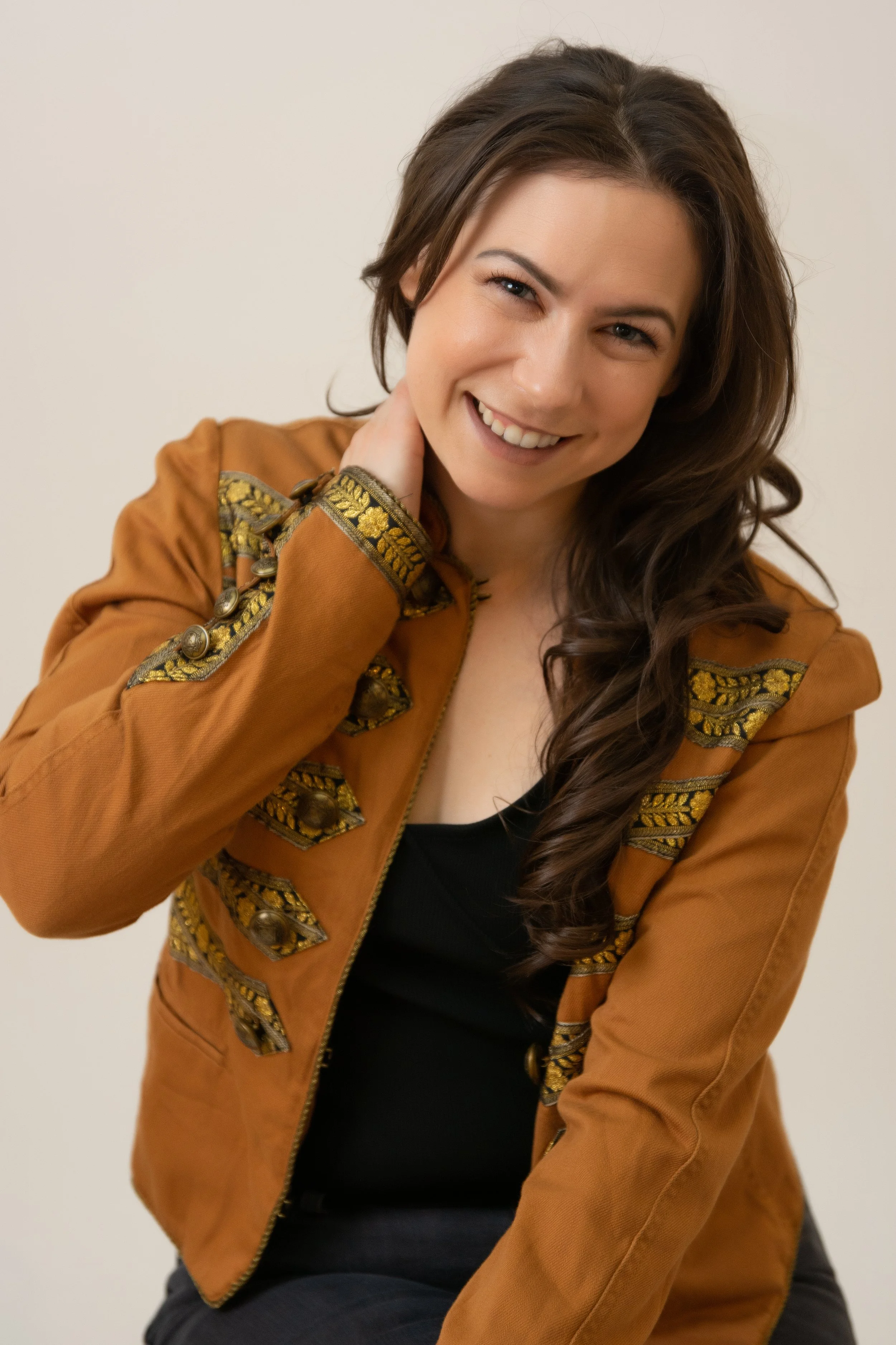A woman with long brown hair, smiling, wearing a brown jacket with decorative gold and black embroidery, sitting against a plain white background.
