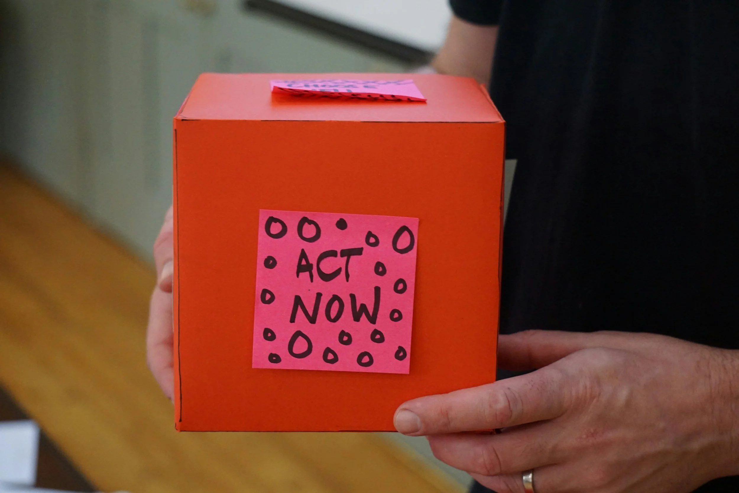Close-up of a person holding an orange box with a pink sticker that says 'ACT NOW!' in black handwritten letters, decorated with black circles.