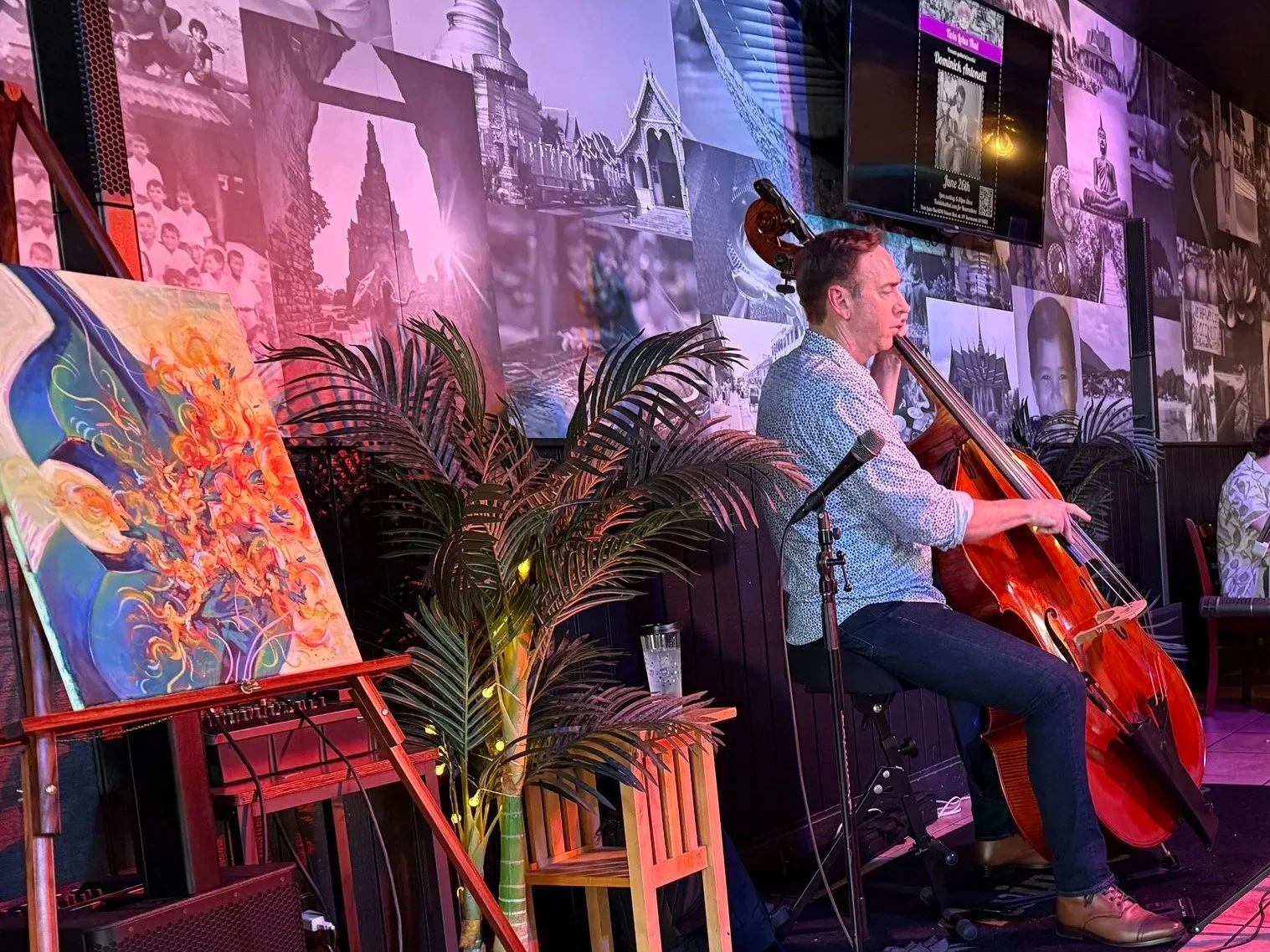 A man playing a cello at a restaurant during a live music performance. The restaurant has black and white photographs on the wall and colorful artwork, with diners seated at tables watching the musician.