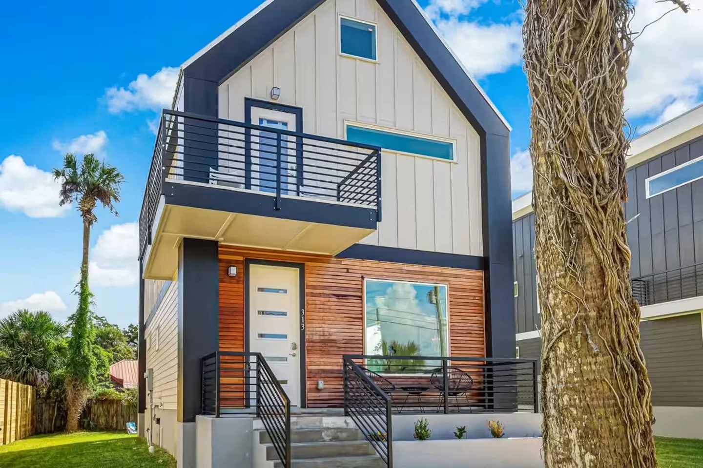 Modern two-story house with a balcony, white and wood exterior, black railings, large window, and a small front porch with steps, surrounded by palm trees and greenery.