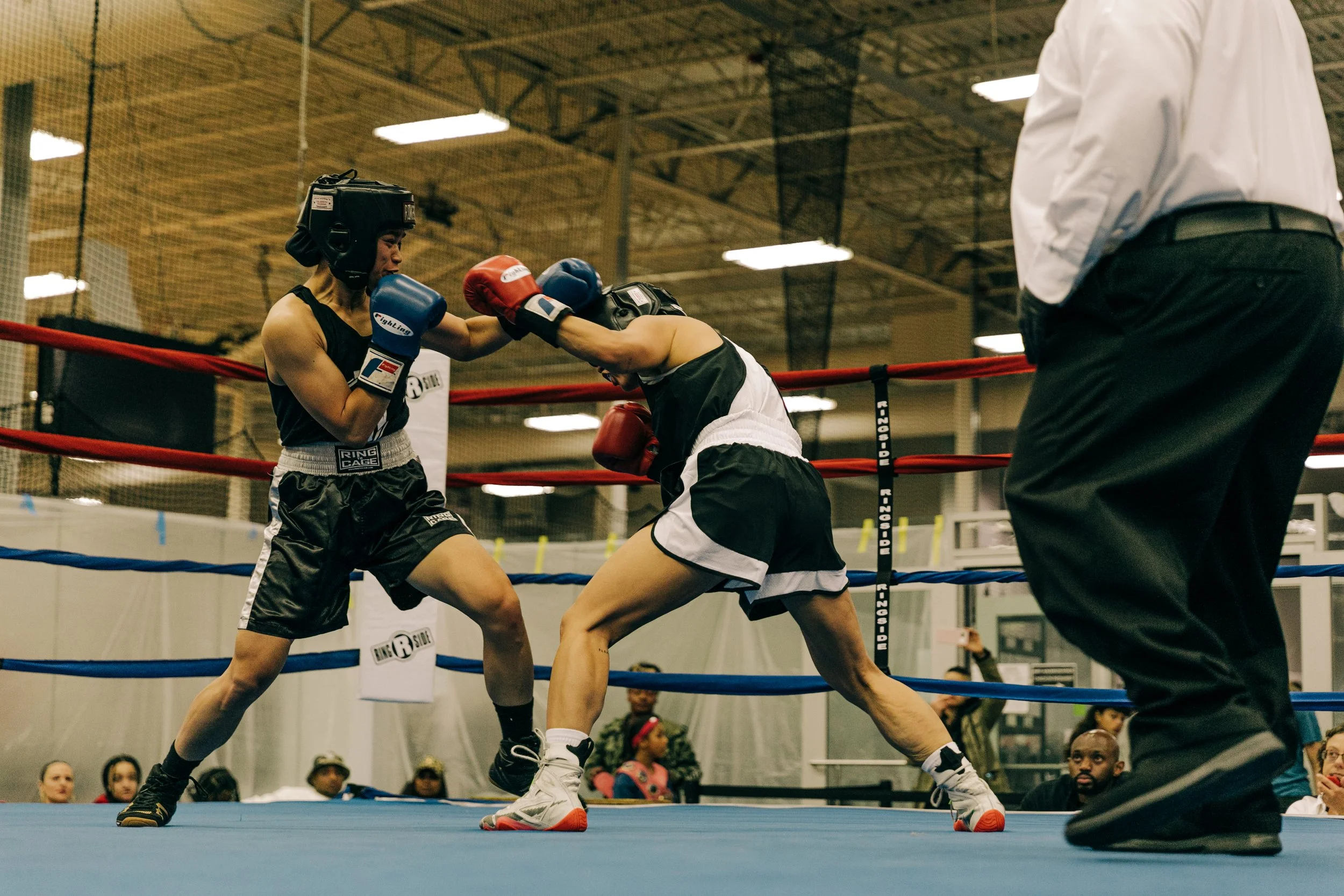 Two women boxing in a ring with a referee watching, and an audience in the background.