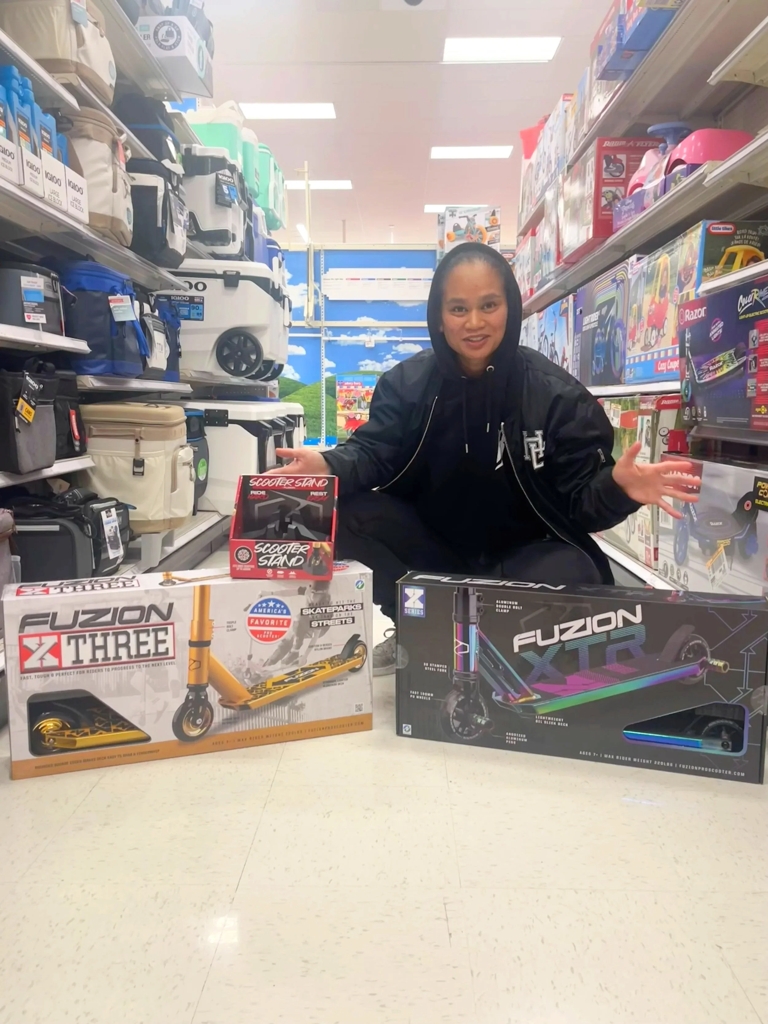 Jackie crouching in an aisle of a store, presenting boxed toy scooters on display shelves. The boxes show images of scooters and colorful graphics.