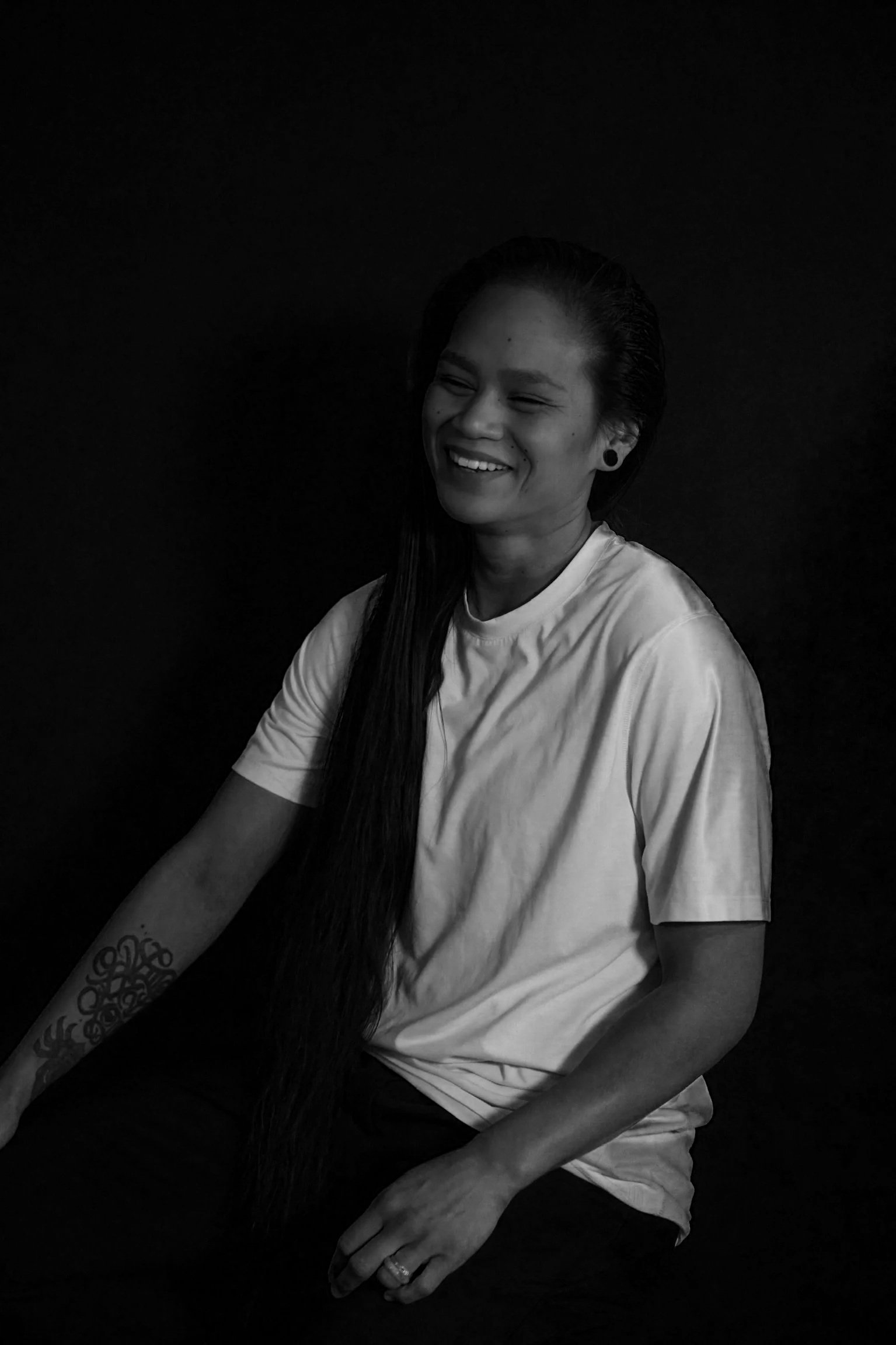 Black and white photo of a woman with long hair, smiling and sitting against a dark background. She has a tattoo on her left forearm, earrings, and a ring on her left hand.