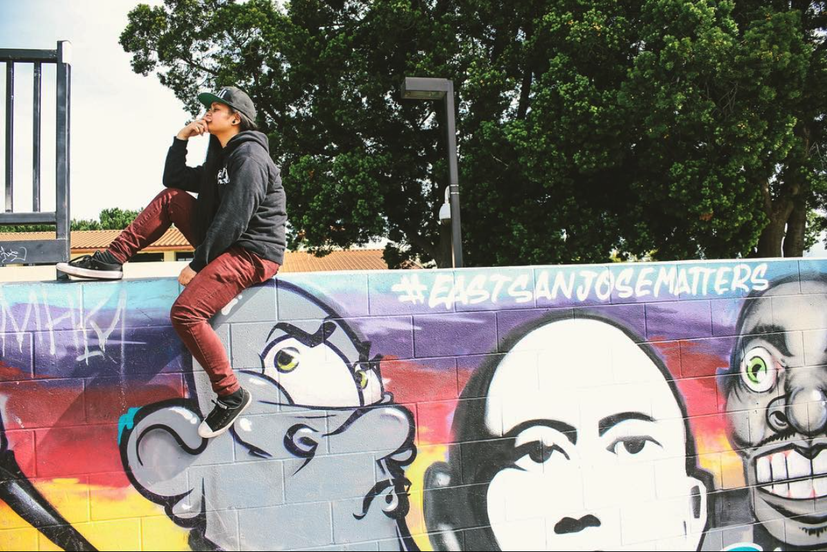 A person wearing a black hoodie, red pants, and a baseball cap sitting on a colorful mural wall with graffiti art, trees, and a building in the background.