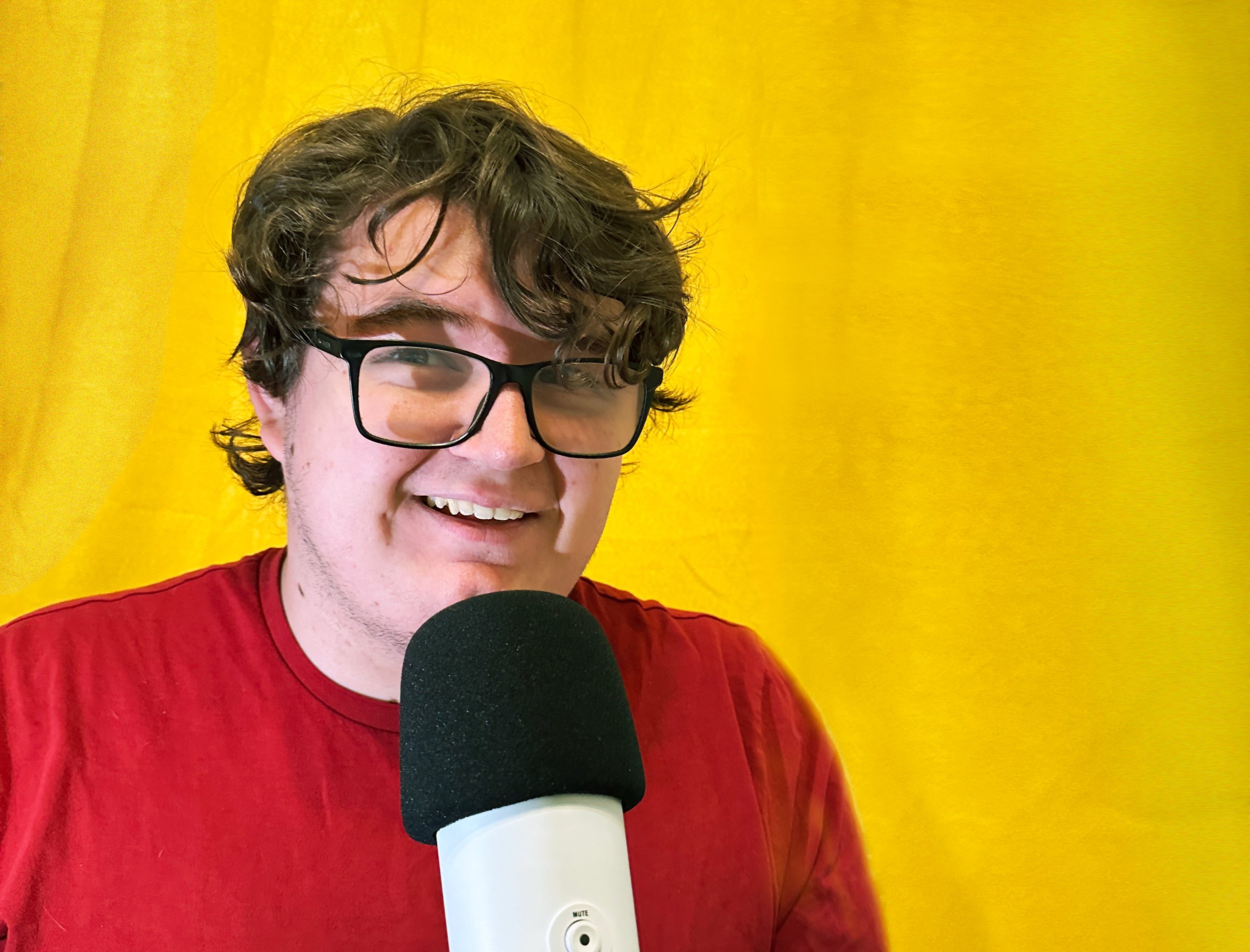 A person wearing glasses and a red shirt smiling at the camera in front of a yellow backdrop, holding a white and black microphone.
