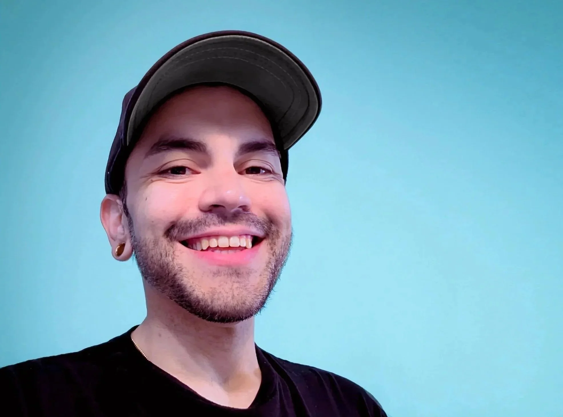 Close-up of a smiling man with earrings, wearing a black shirt and a gray baseball cap, against a light blue background.