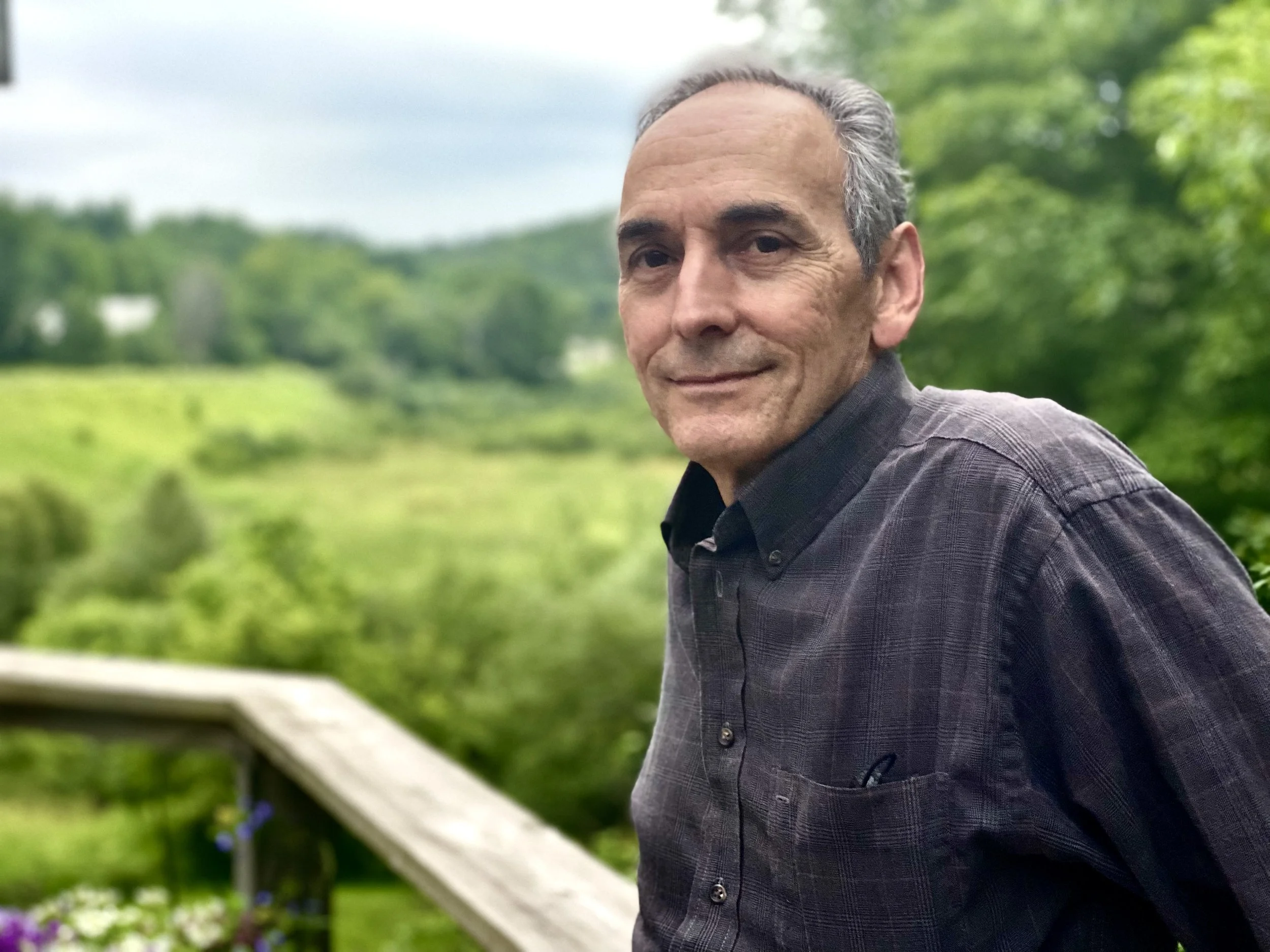A middle-aged man with gray hair and a slight smile, wearing a dark shirt, stands on a wooden deck with green trees in the background.
